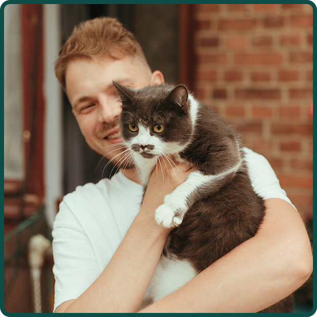 A young man is smiling and holding a black and white cat outside in front of a brick building.
