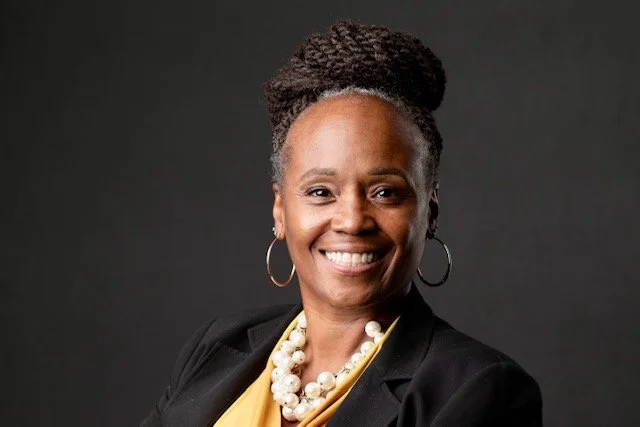 Portrait of a smiling Black woman with braided hair styled in an updo, wearing a black blazer, a yellow blouse, a pearl necklace, and hoop earrings, against a dark background.
