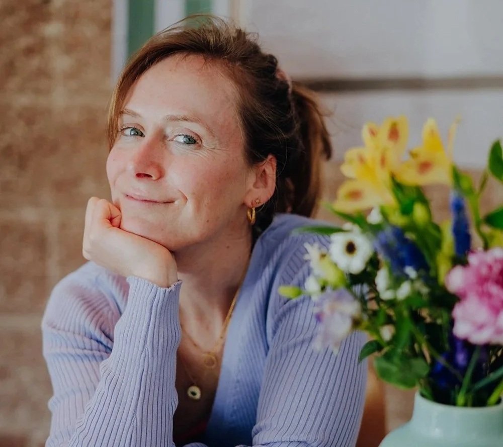 Eine Frau mit dunkelblonden Haaren, lächelnd, sitzt an einem Tisch neben einem Blumenstrauß mit gelben, weißen, blauen und pinken Blumen.