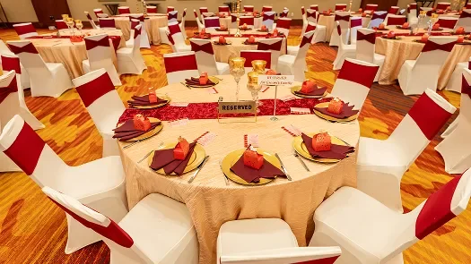 Banquet hall with round tables set for an event, decorated with red and gold accents, and chairs with red and white covers.