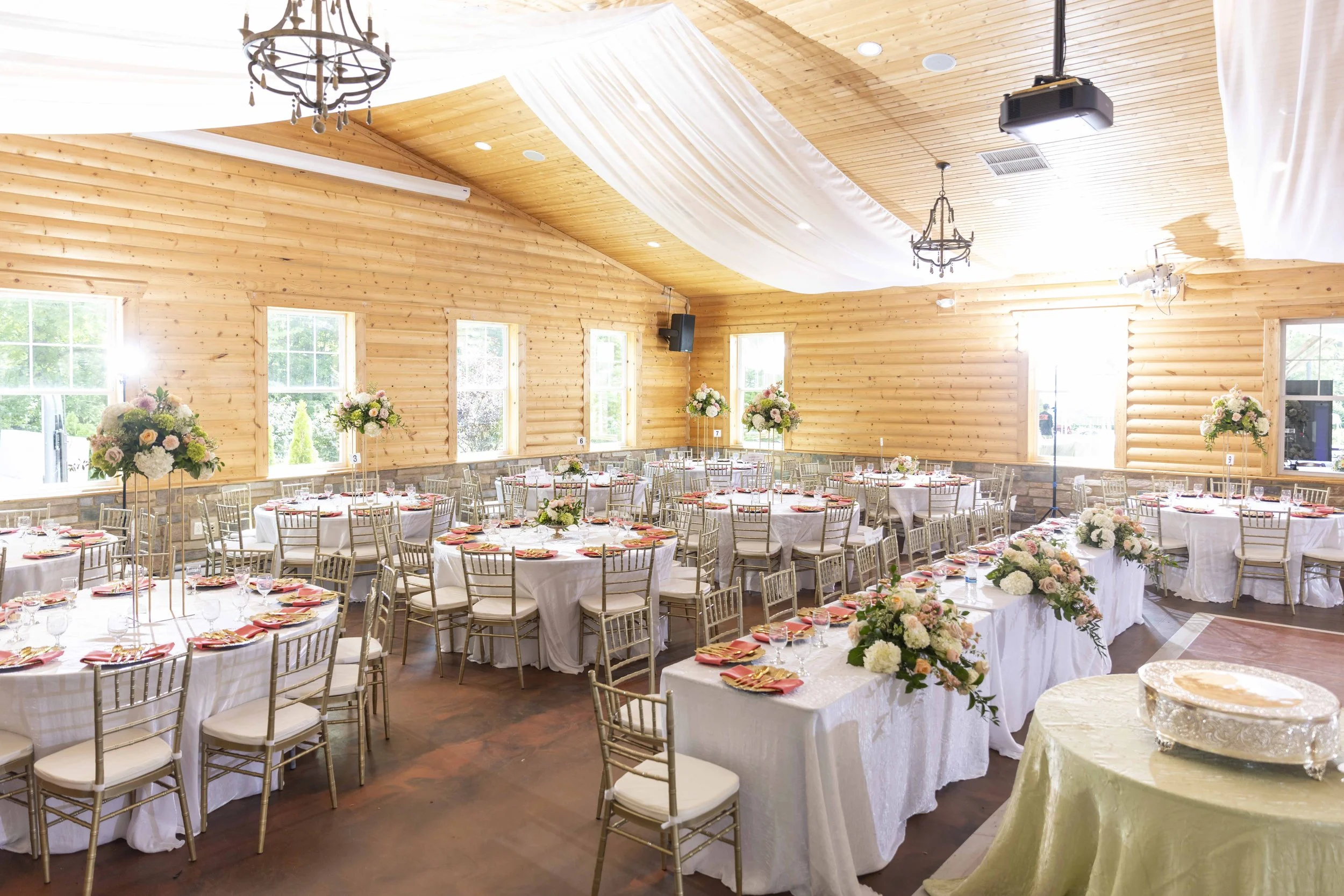 Elegant banquet hall with round tables covered in white tablecloths, decorated with floral centerpieces, set with plates, glasses, and napkins. The room has wooden walls, high windows, chandeliers, and a ceiling draped with white fabric for a wedding or special event.