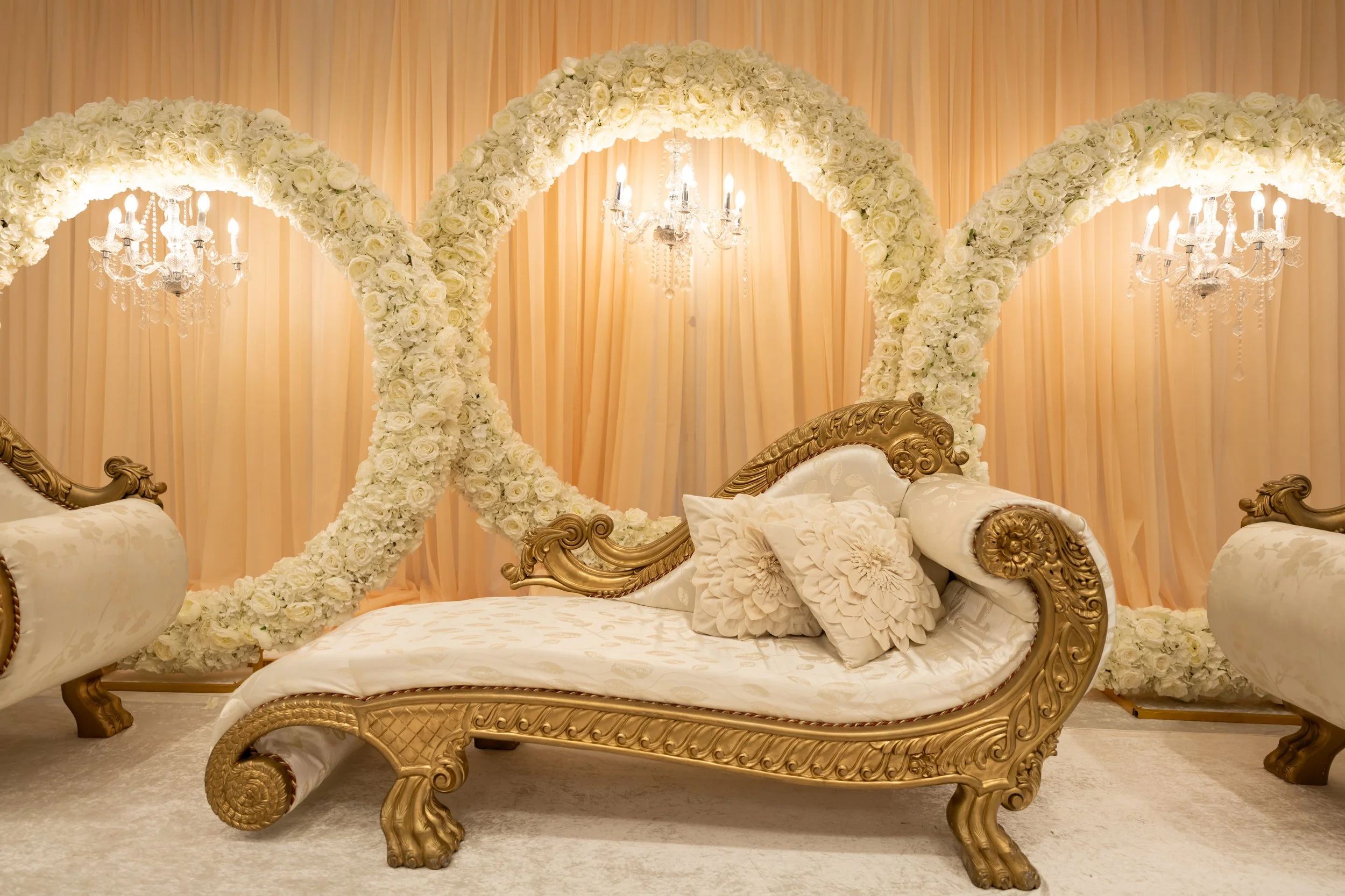 A luxurious wedding bed with an ornate gold frame, decorated with white floral arrangements, with cream-colored cushions. Behind the bed, there are large floral arches and chandeliers hanging from the ceiling, creating an elegant backdrop.