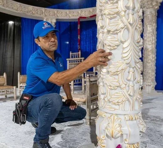 A man in a blue polo shirt and cap examines a large ornate white column with gold accents in an indoor setting with blue curtains and chairs.