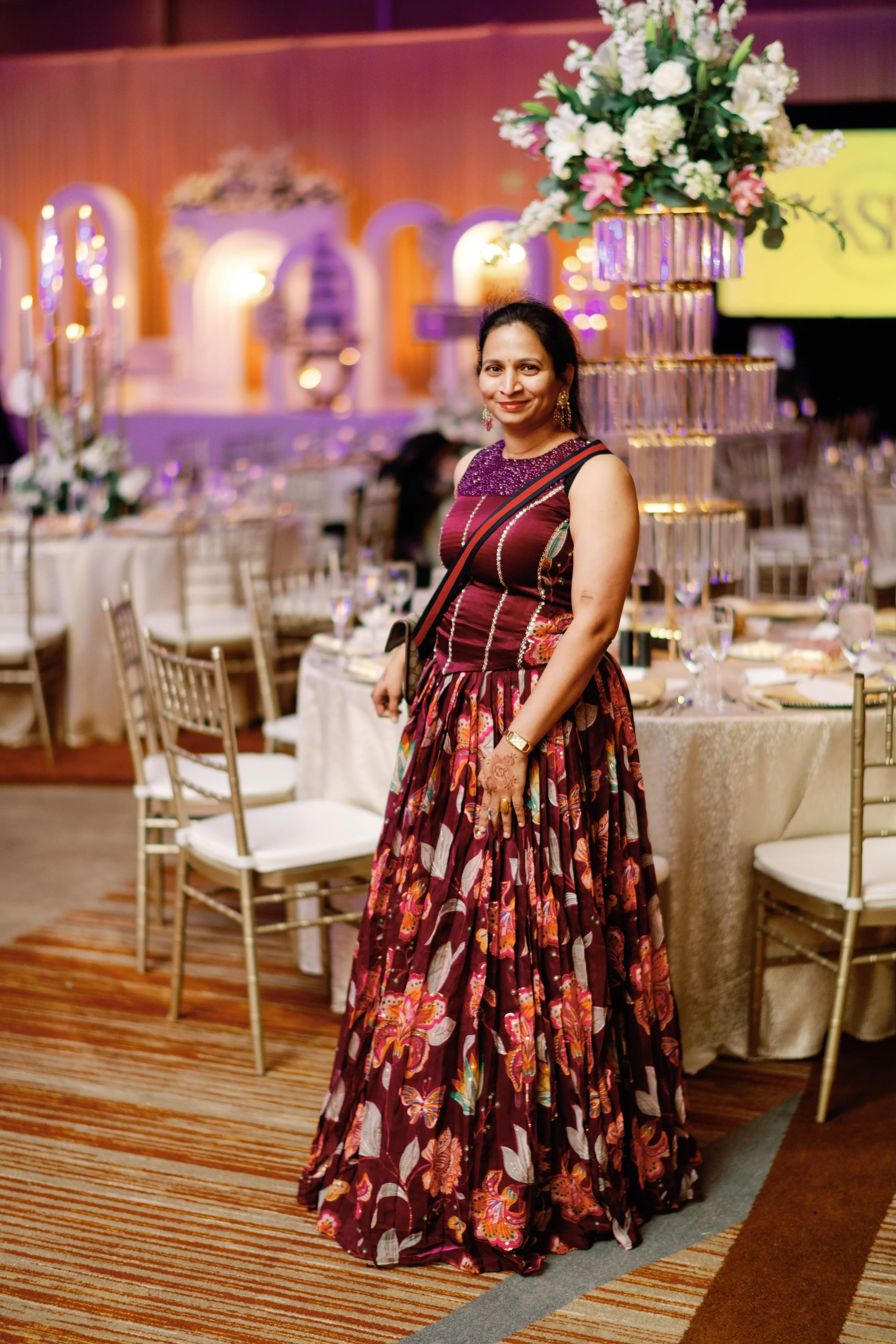 A woman wearing a colorful traditional dress standing in a decorated banquet hall with round tables, floral centerpieces, and elegant lighting for a celebration or wedding event.