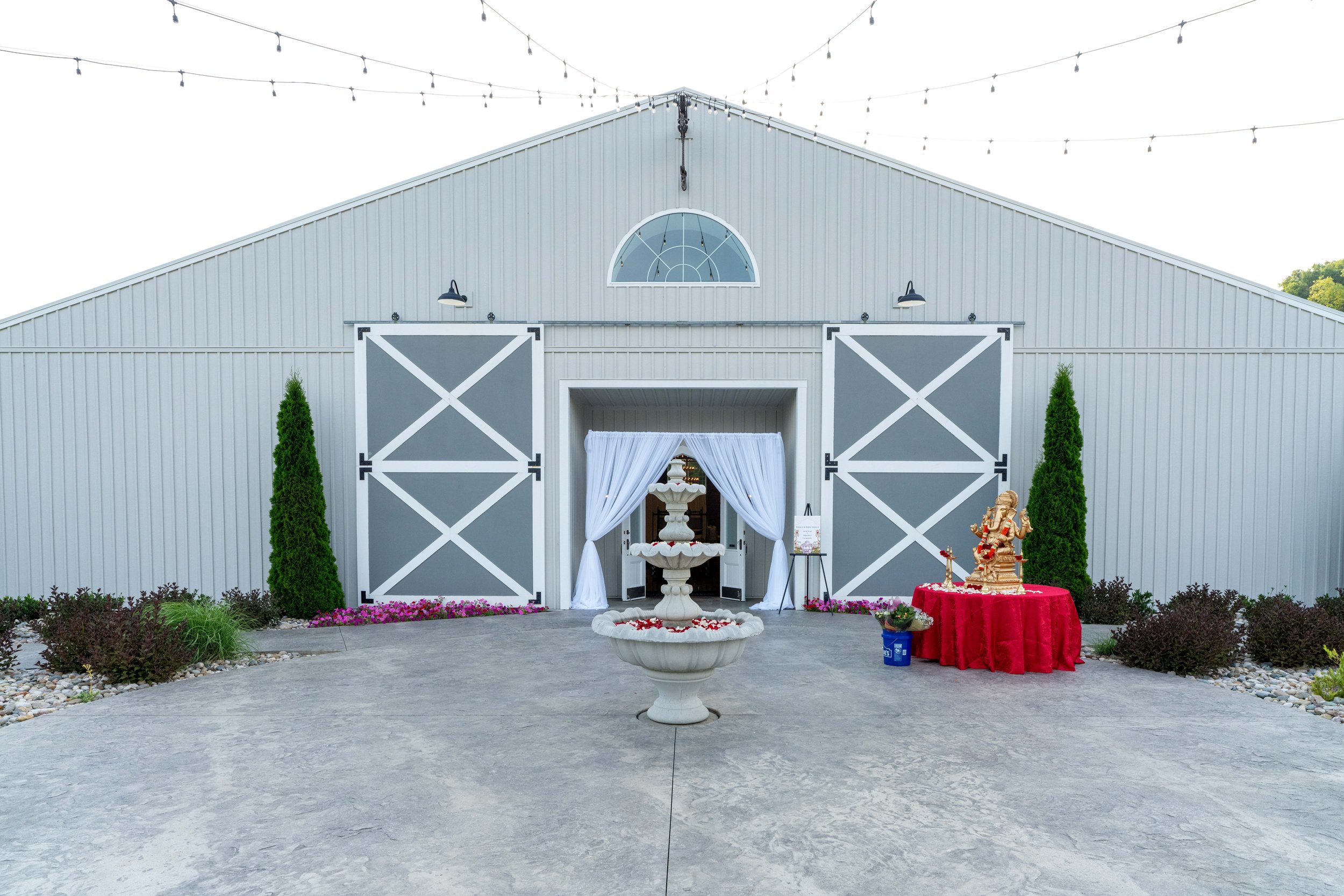 A white barn decorated for a celebration, with open sliding doors, a white draped archway, a table with a golden Ganesh statue, and string lights overhead.