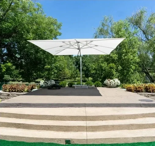 An outdoor stage with steps, a white patio umbrella, and potted plants, surrounded by green trees and a clear blue sky.
