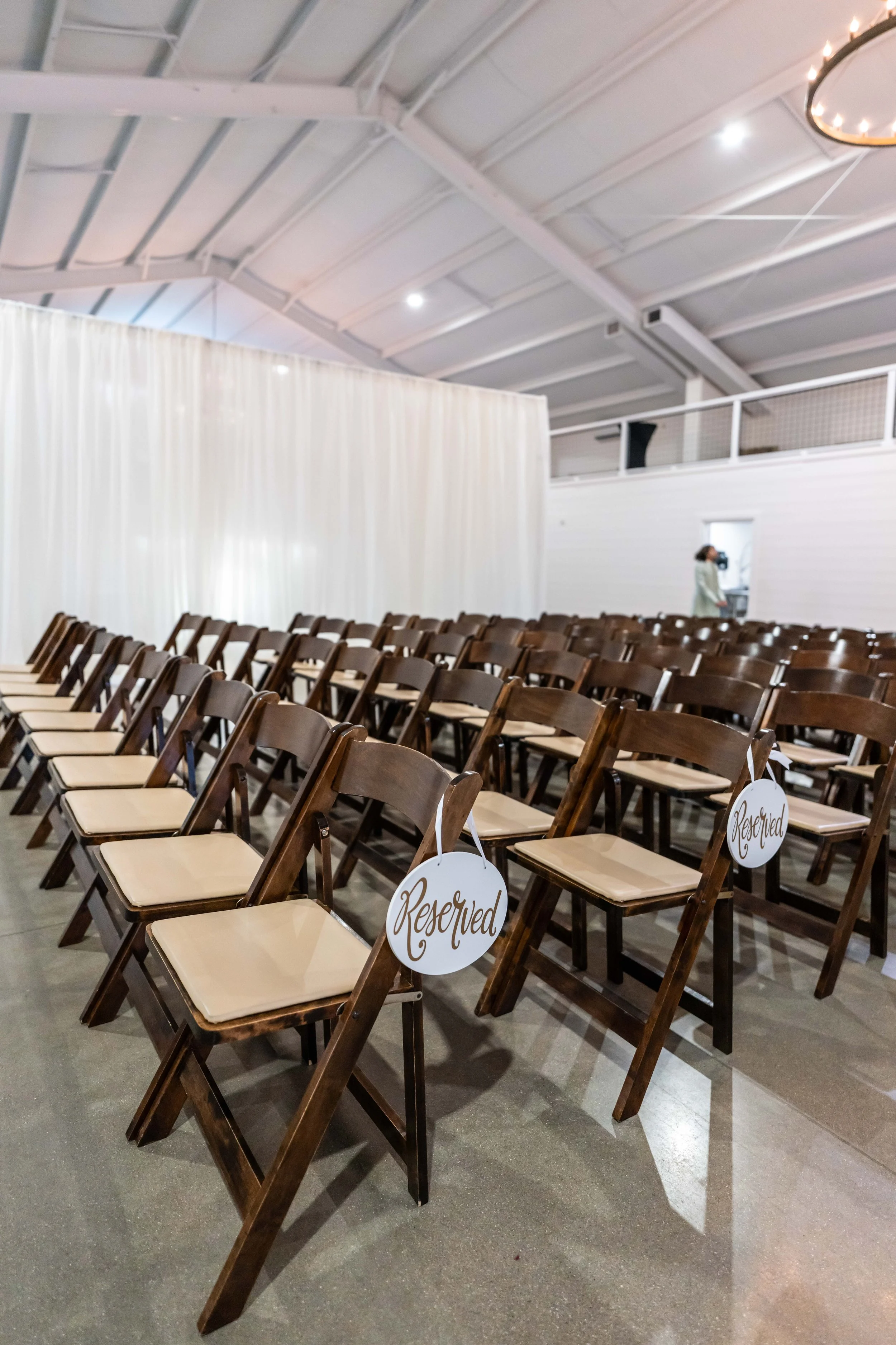 Rows of brown wooden chairs with beige cushions, some with 'Reserved' signs hanging on them, in an indoor event space with high ceilings, white walls, and a partially visible circular chandelier.