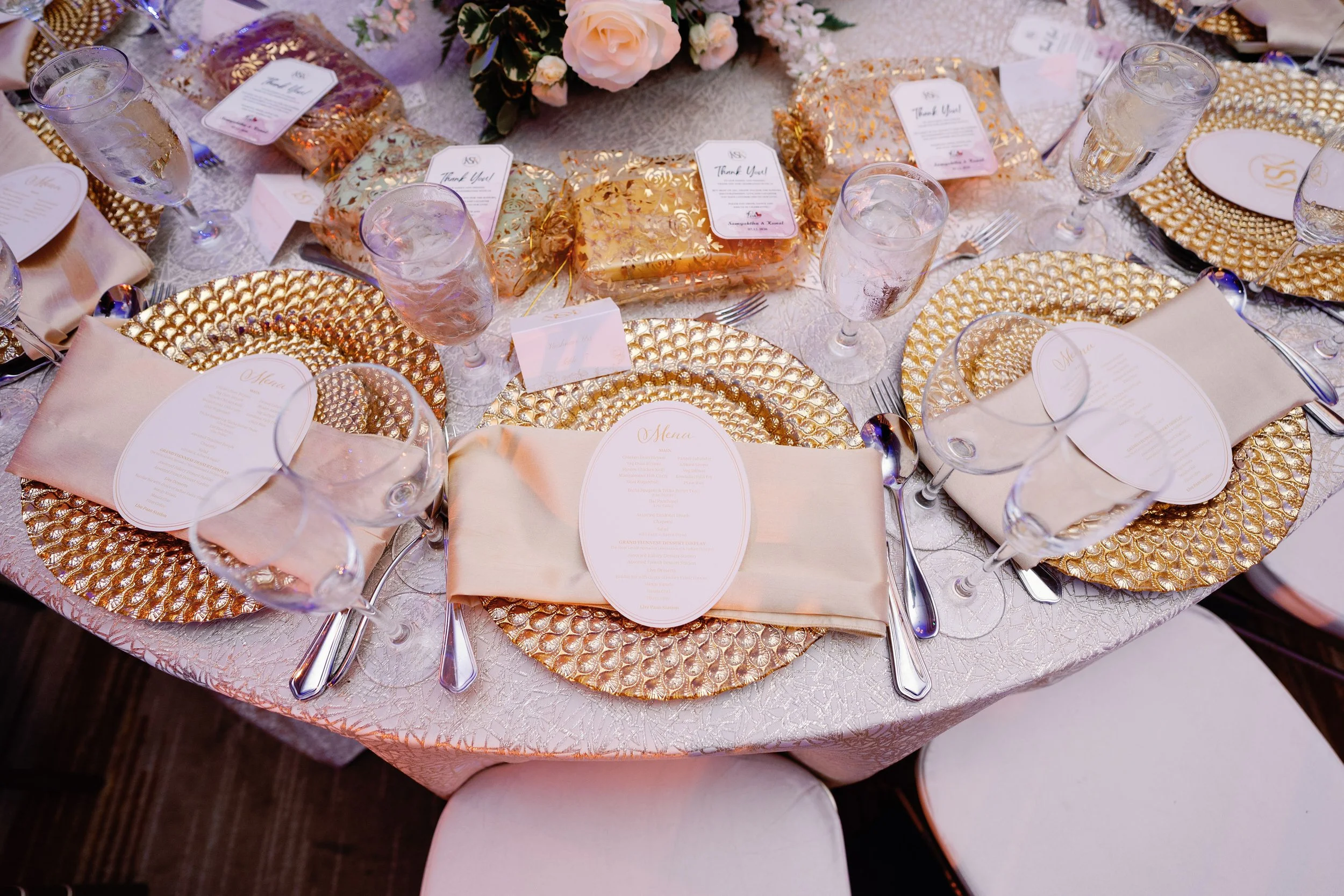 A formal banquet table setting with gold-colored placemats, printed menus, glassware filled with ice water, cream-colored napkins, and decorative wrapped party favors or gifts at the center, with a floral centerpiece in pink and white.