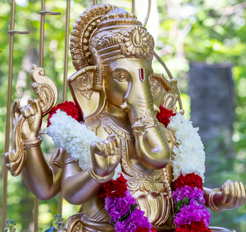 Golden statue of Lord Ganesha adorned with flower garlands, set outdoors with greenery in the background.
