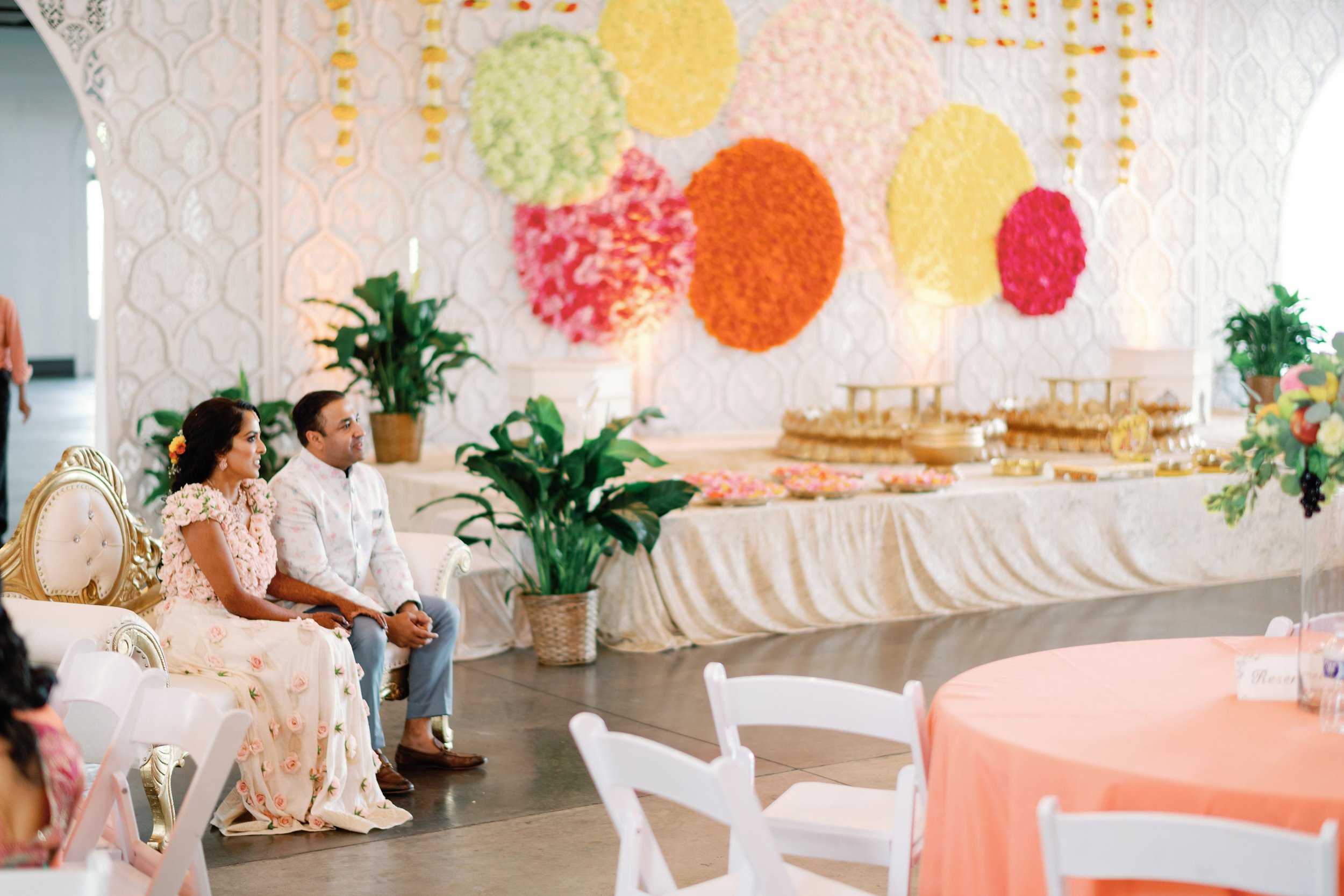 A decorated banquet hall with a floral wall backdrop, featuring orange, pink, yellow, and green pom-poms. Guests dressed in traditional attire are seated on ornate white and gold chairs in front of a table with orange and pink decorations and floral arrangements.
