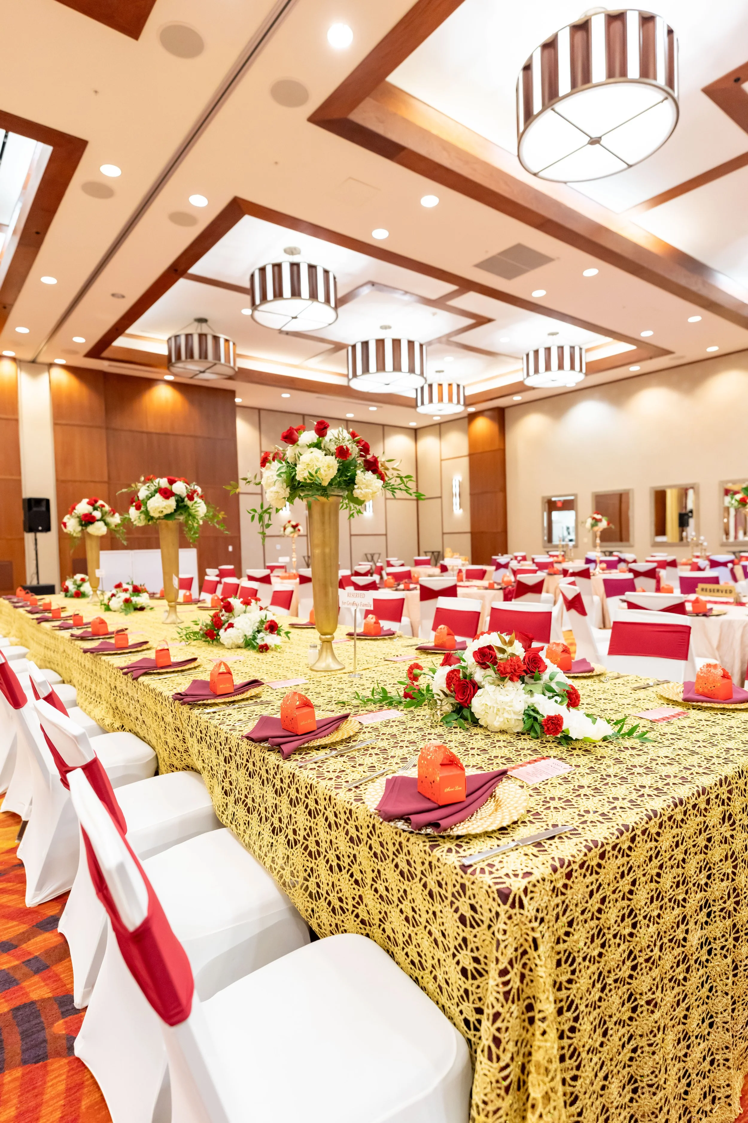 Elegant banquet hall decorated for a celebration with long gold-patterned tablecloths, floral centerpieces, and chairs draped with white covers and red sashes. The room features wooden accents, circular ceiling lights, and a warm lighting ambiance.