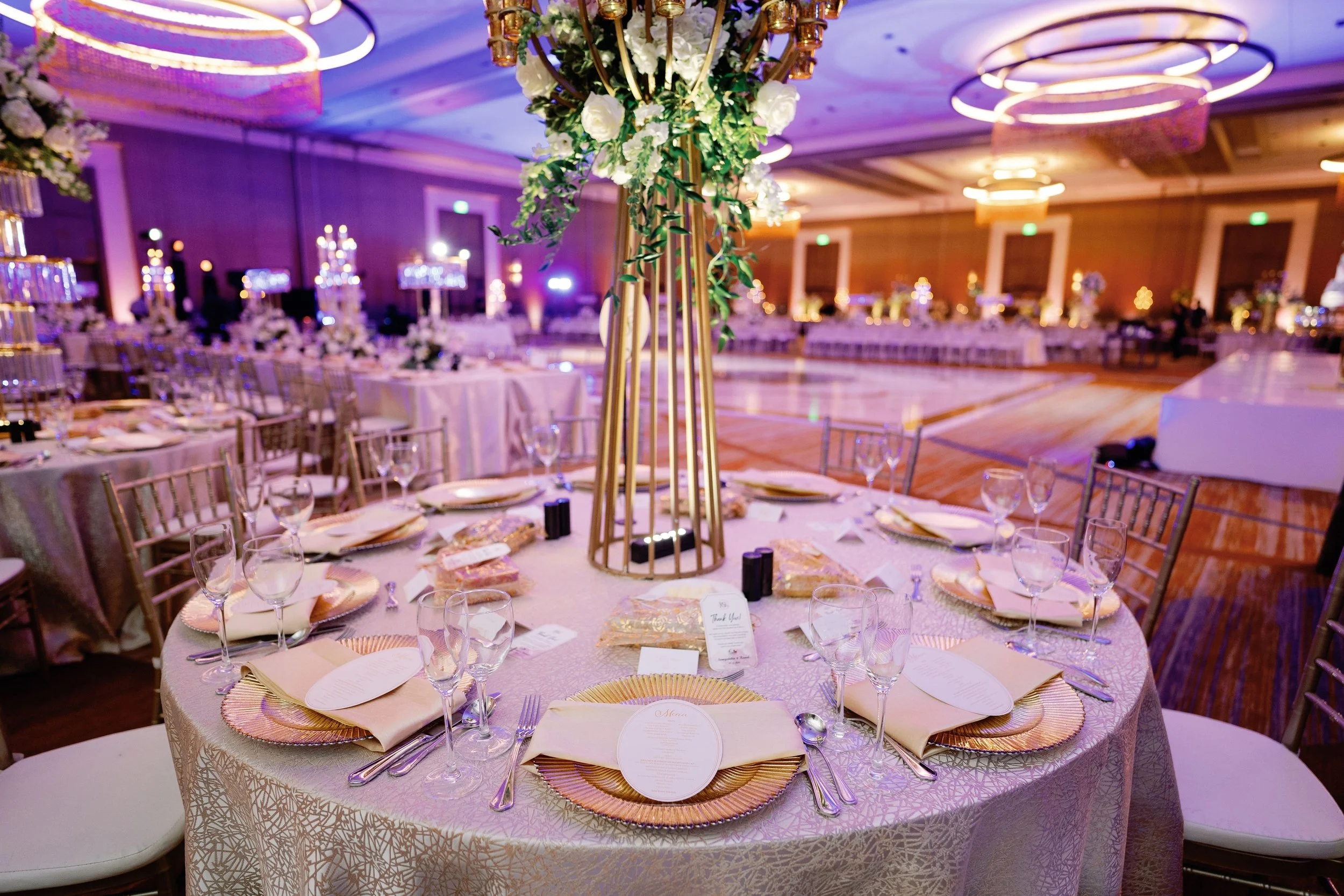 Elegant banquet hall decorated for a wedding or special event with round tables covered in white tablecloths, set with glassware, plates, and gold chargers, and adorned with tall floral centerpieces featuring white flowers and gold accents.
