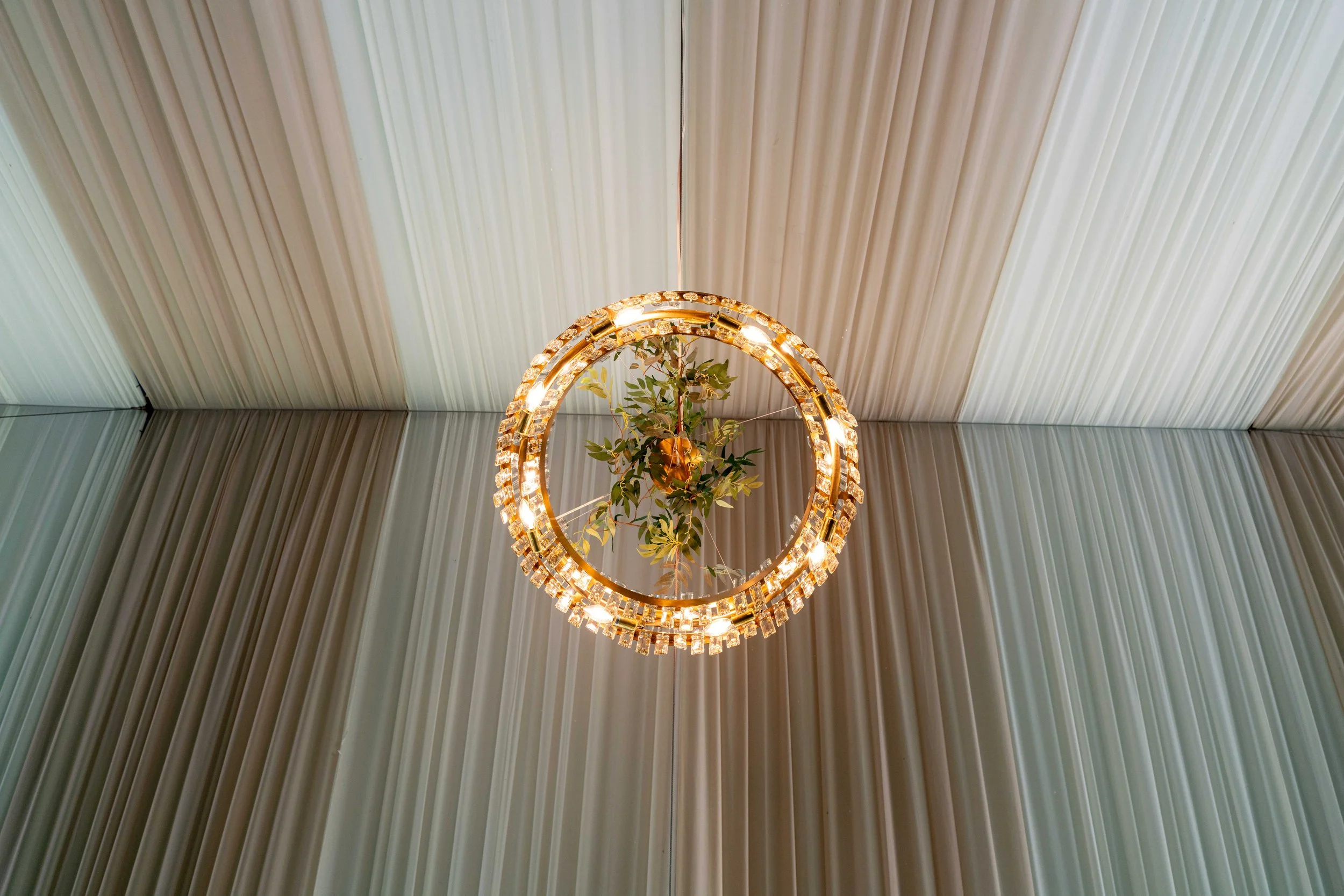 A circular chandelier with lights and a small tree hanging inside, set against a background of vertical pale curtains on the ceiling and walls.