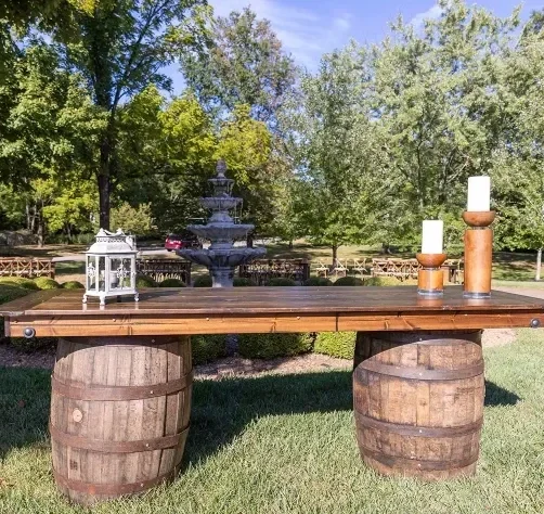 Outdoor table made from two wooden barrels with a wooden top, decorated with candles and a lantern, in a garden with trees and a fountain in the background.