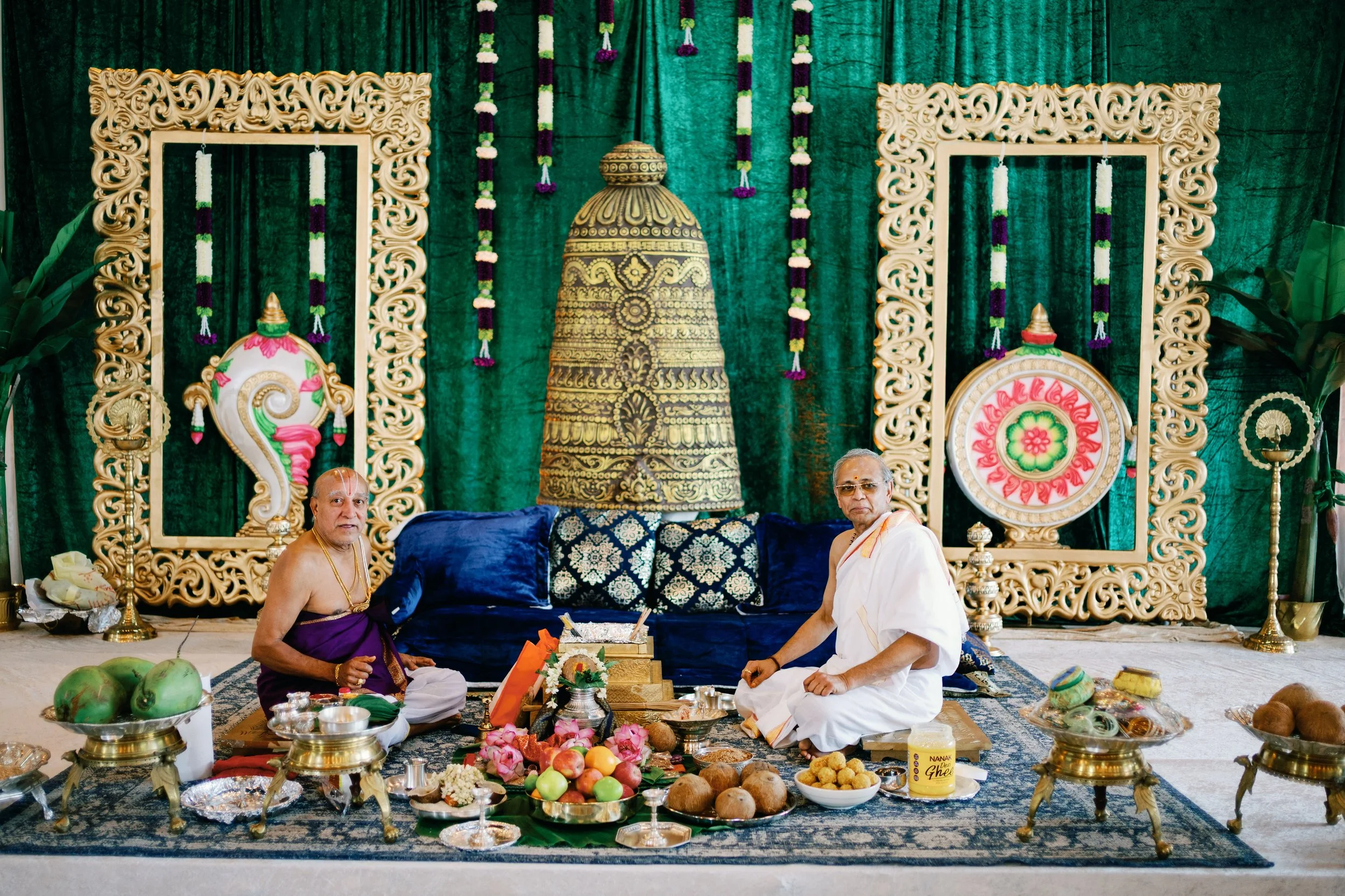 Two men sitting on the floor in front of a decorated altar with religious artifacts, fruits, and offerings. The background features green drapes, ornamental frames, and hanging flower garlands.