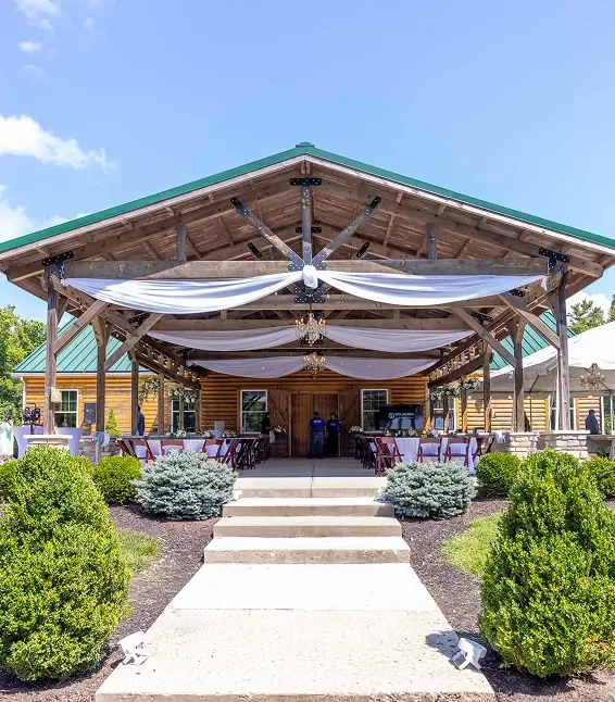Outdoor event tent with white drapery, surrounded by green bushes, in front of a wooden building with a green roof.