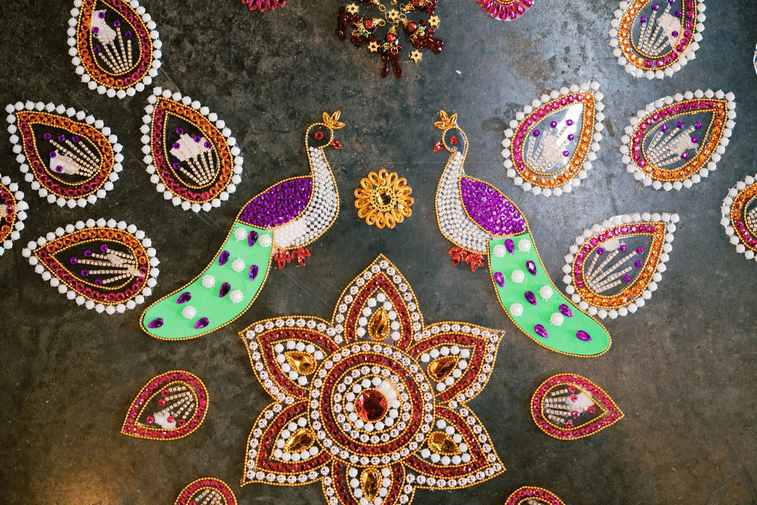 Colorful traditional Indian decorative items, including peacock motifs and floral designs made with beads and stones, arranged on a dark surface.