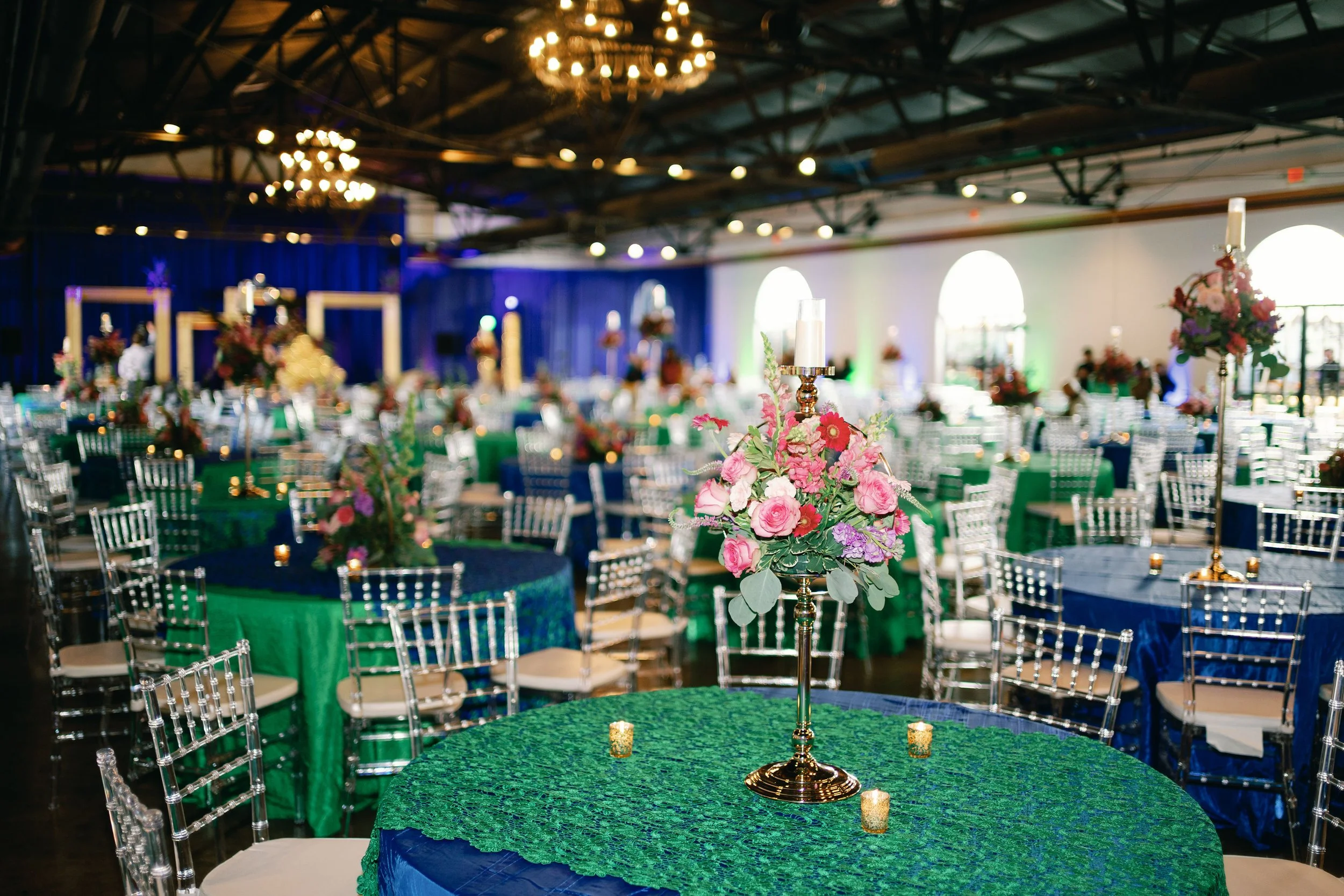 Indoor event space decorated with green and blue tablecloths, floral centerpieces with pink and purple flowers, and candle holders. Chandeliers hang from the ceiling, and they are illuminated. The setting appears festive, possibly for a wedding or banquet.