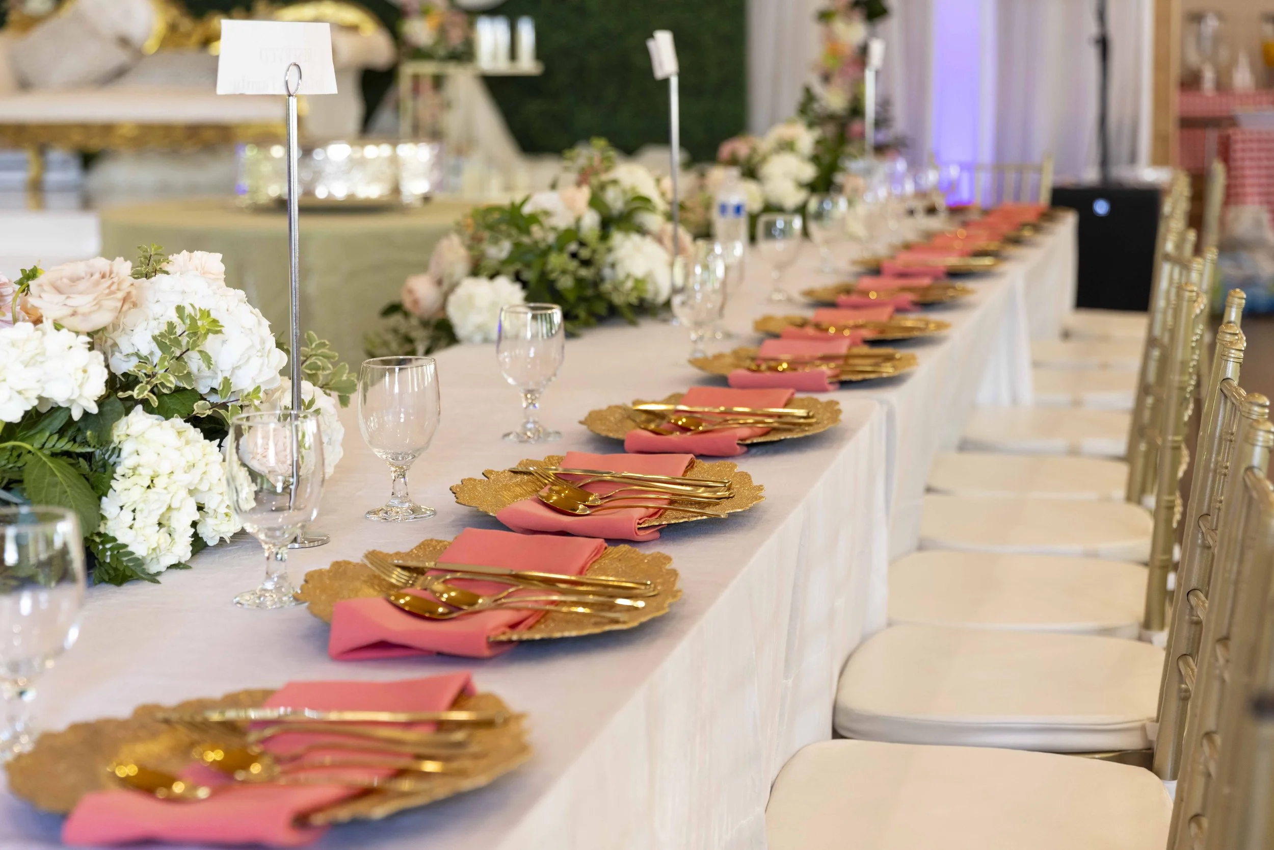 A long banquet table set for a formal event with pink napkins, gold utensils, and floral centerpieces of white and pink flowers. Gold chairs line the table, and there are signs on stands and glasses set for each place.