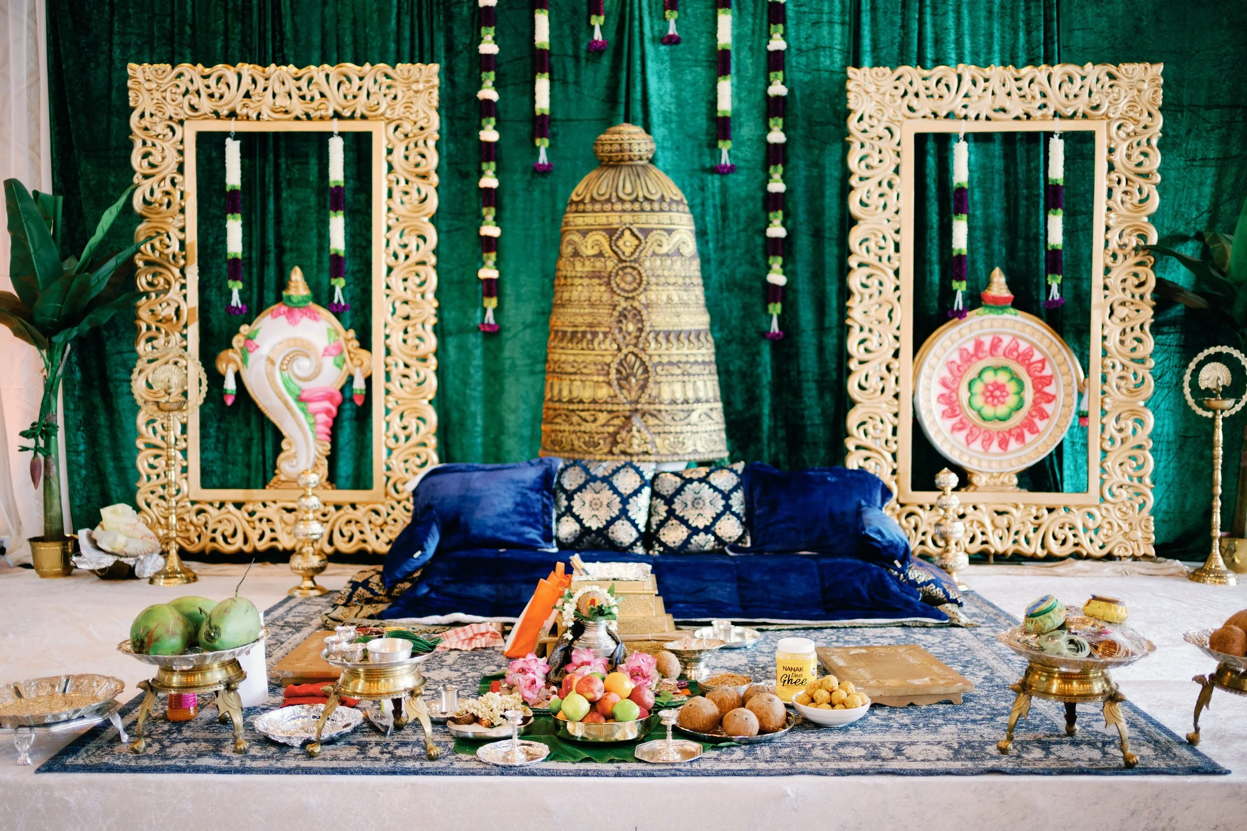 A traditional Indian religious ceremony setup with a decorative backdrop, ornate frames, hanging floral garlands, a large decorated bell or temple centerpiece, and various offerings including fruits, sweets, and coconut on a decorated platform.
