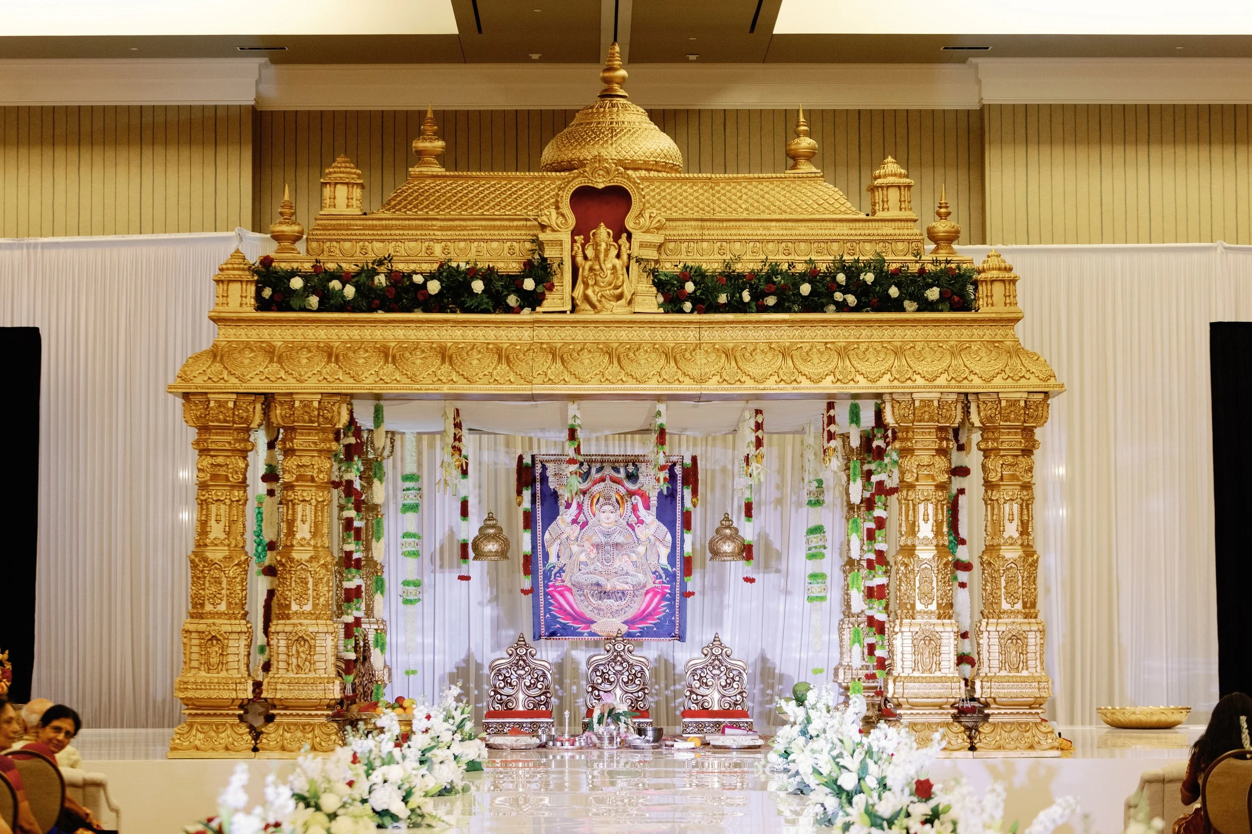 A large ornate gold temple structure decorated with flowers, with three chairs and a colorful image of a deity in the background, in an indoor setting.
