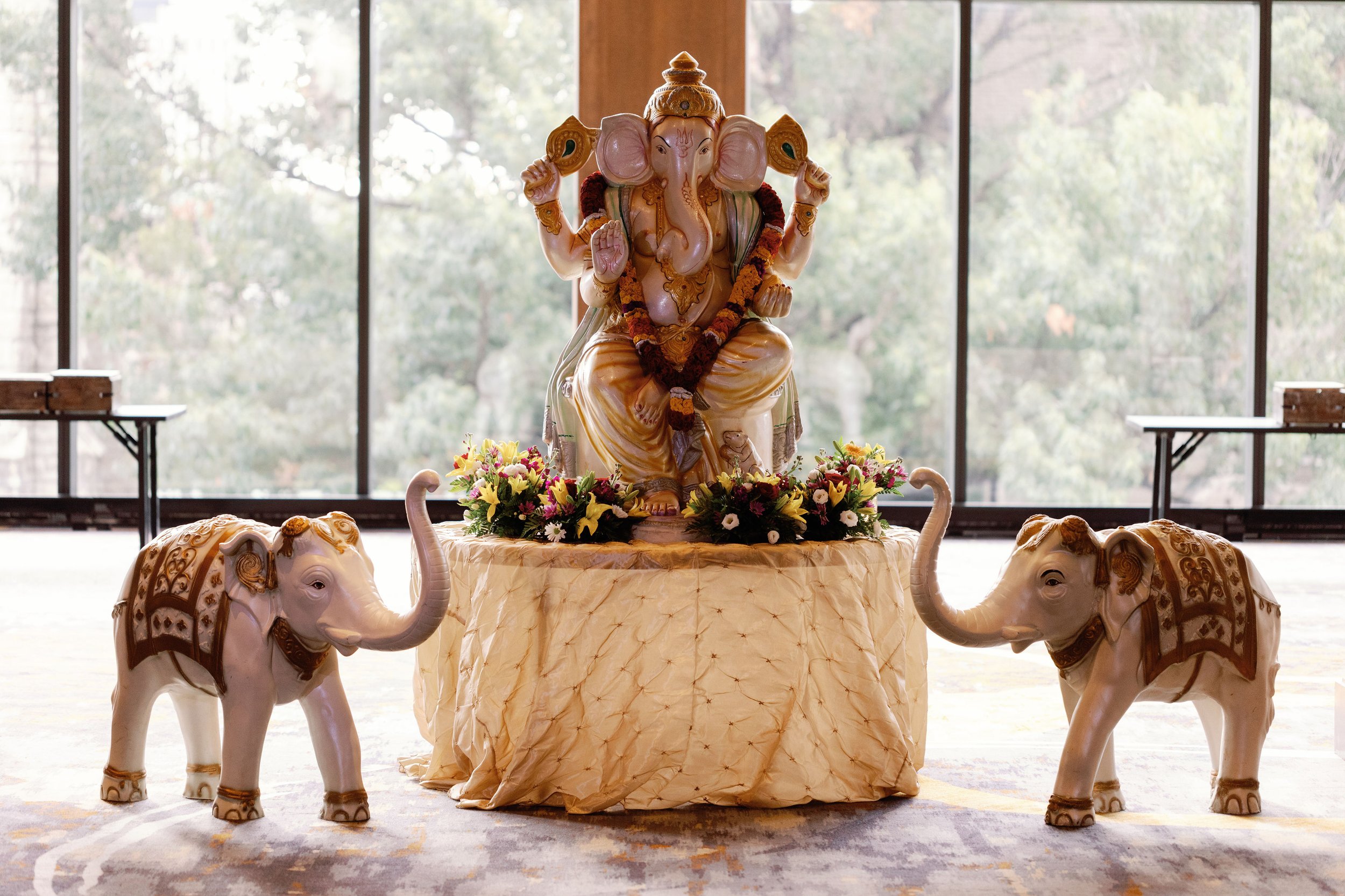A decorative sculpture of the Hindu deity Ganesha seated on a table, surrounded by three elephant-shaped statues, with a backdrop of large windows and natural scenery.