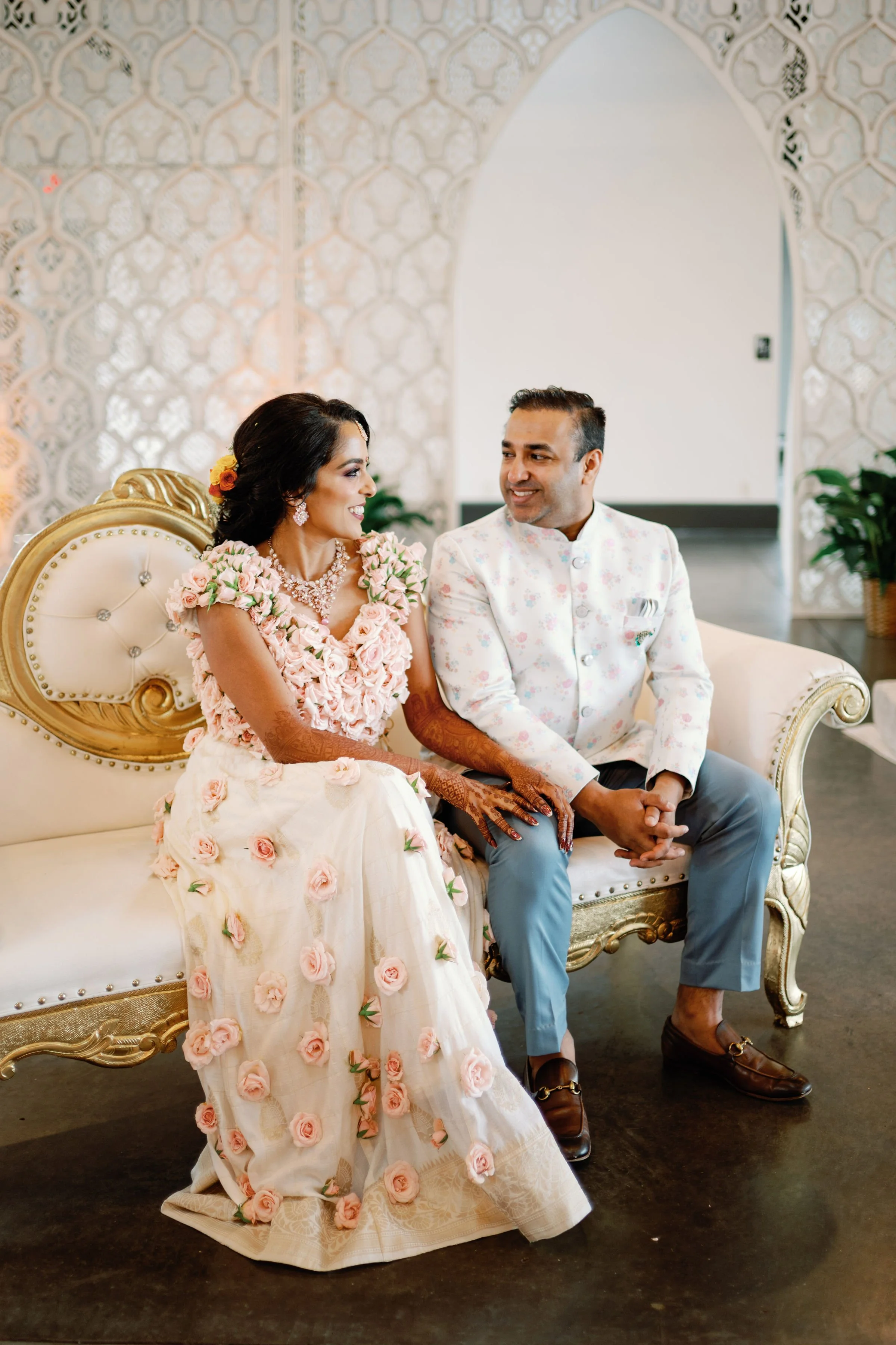 A bride and groom sitting on a vintage-style cream and gold sofa at their wedding. The bride wears a floral dress with pink roses and intricate jewelry. The groom wears a white floral blazer, gray pants, and brown loafers. They are smiling and looking at each other against a decorative white wall with plants nearby.