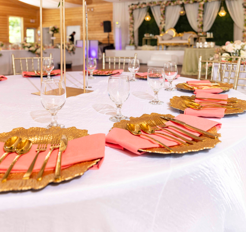 Elegant banquet table set with pink napkins, gold cutlery, and glassware, decorated for a celebration with a decorated stage in the background.