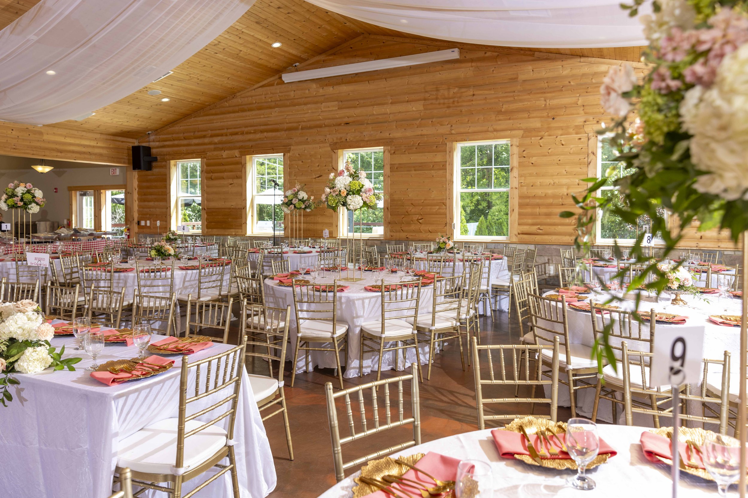 Indoor wedding reception with round tables covered in white tablecloths, decorated with pink and white floral centerpieces, gold chairs, and place settings. Large windows revealing green trees outside, and warm wooden walls and ceiling interior.