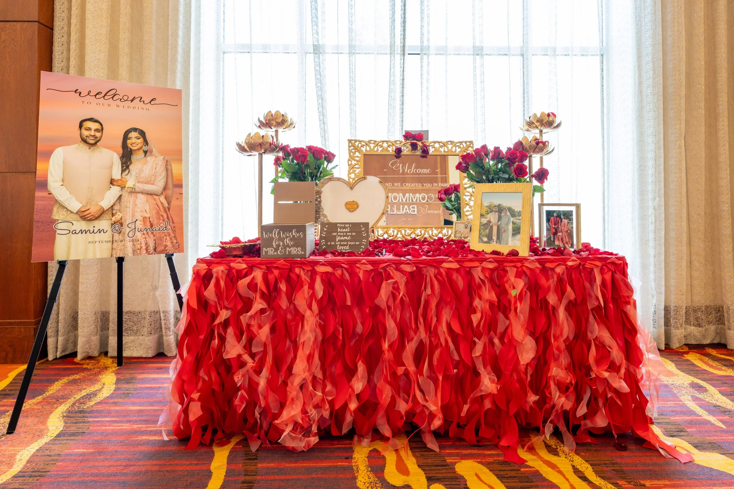 A wedding display table with red and pink roses, framed photos of the couple, greeting sign, and decorative elements, set in front of sheer curtains with a welcome sign to Samin and Junaid.