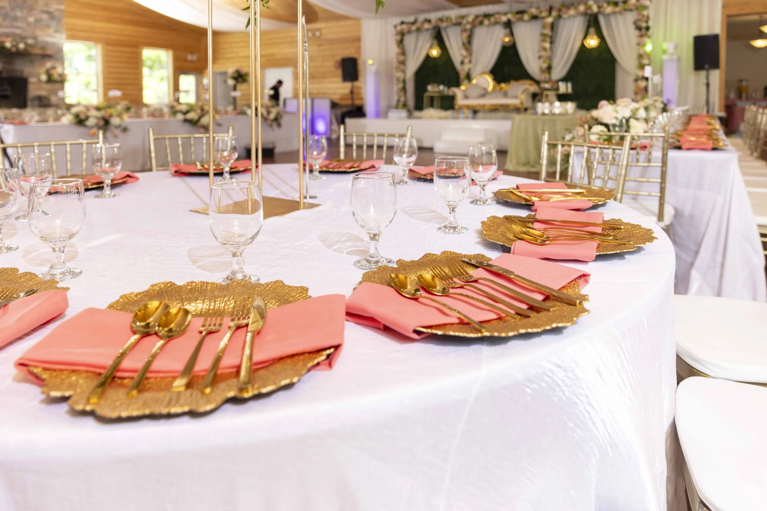 A decorated banquet table set with gold and pink napkins, gold cutlery, and glassware, in a reception hall with floral and elegant decor.