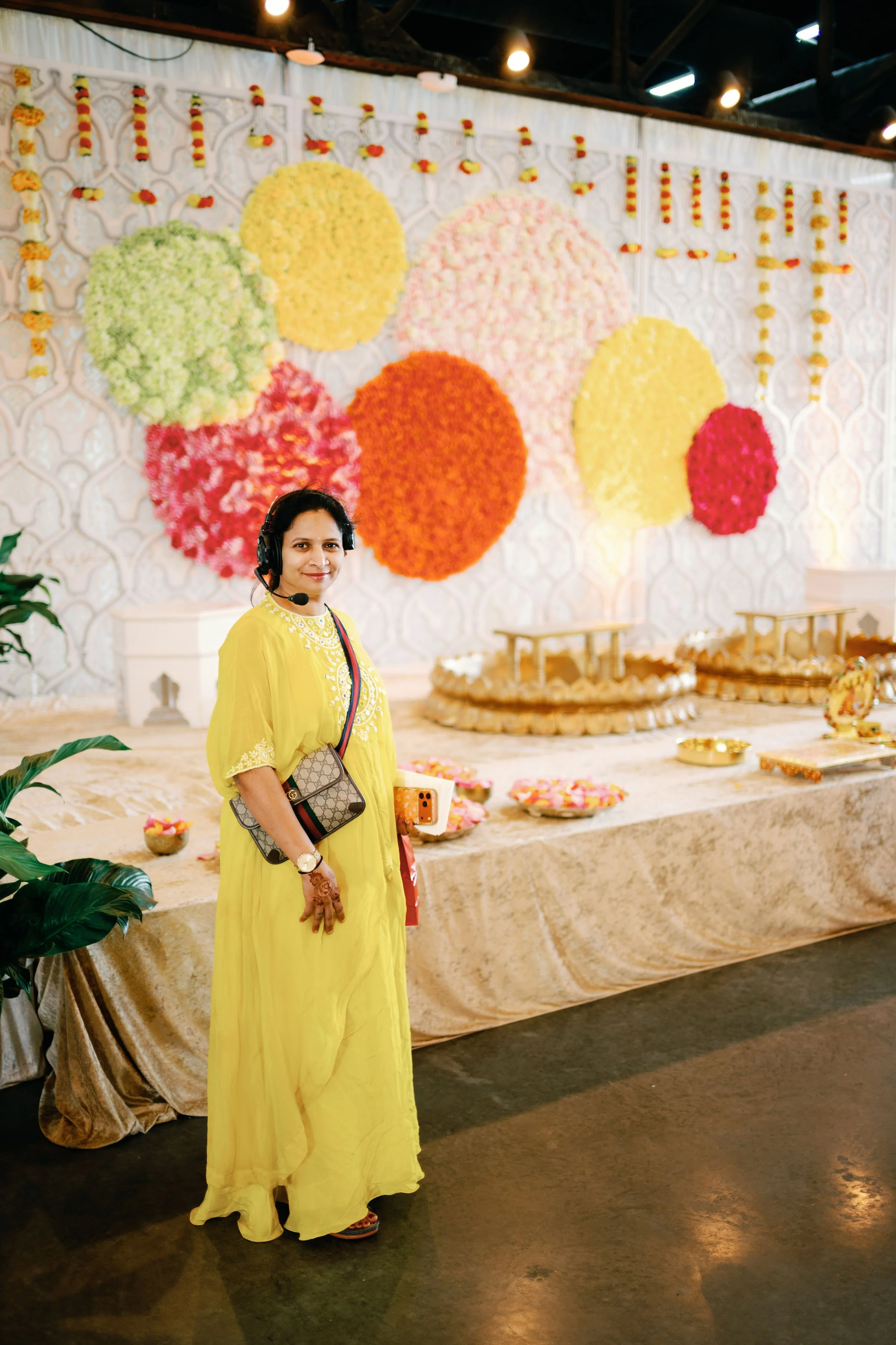 A woman wearing a yellow traditional dress with henna on her hands stands in front of a decorated backdrop with large, colorful floral arrangements, and a table with offerings or decorations at what appears to be a celebration or cultural event.