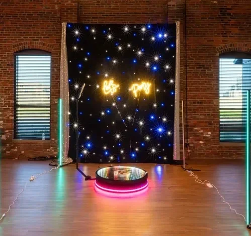 A decorated setup with a starry background, neon sign that says 'Let's Play,' and a circular platform with pink and purple lights in front of a brick wall and two windows.