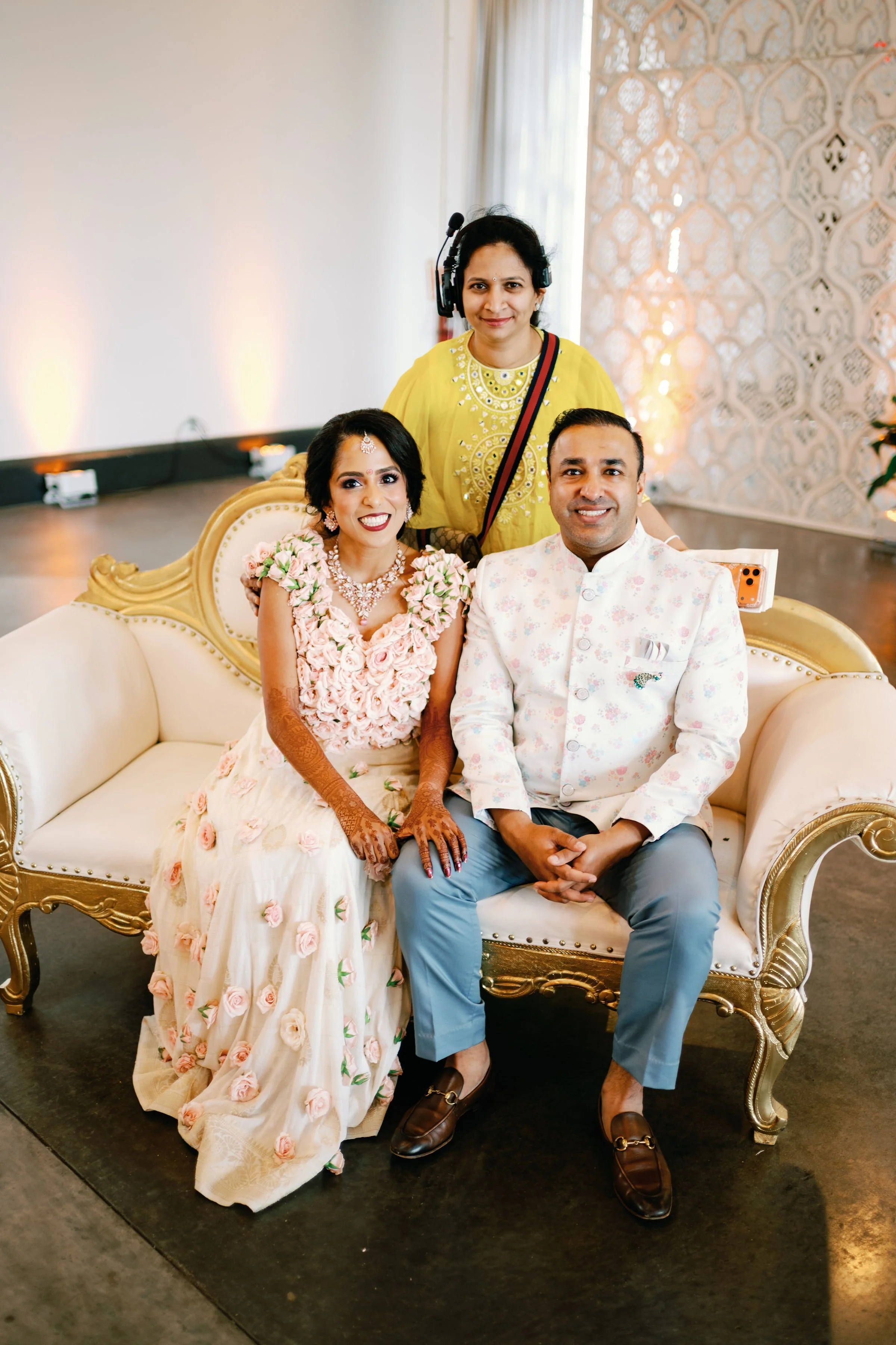 A newlywed couple in traditional Indian wedding attire seated on a white and gold ornate sofa, smiling, with a woman in yellow standing behind them wearing a headset at a wedding reception.
