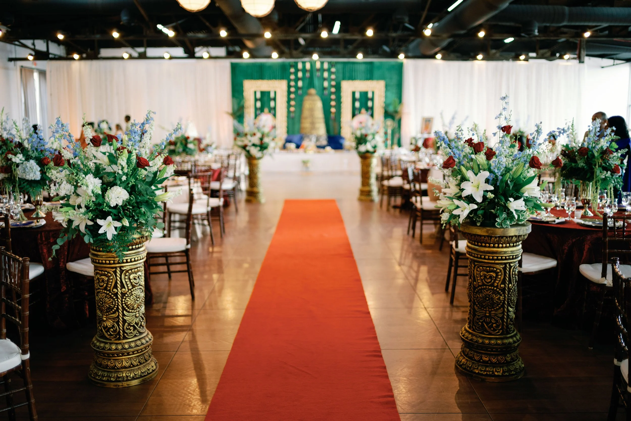 A wedding reception hall decorated with floral arrangements of white lilies, roses, and blue flowers on gold ornate pedestals, with a red carpet leading to a decorated stage with a green backdrop and floral decor, and surrounded by tables and chairs, some of which have people seated.