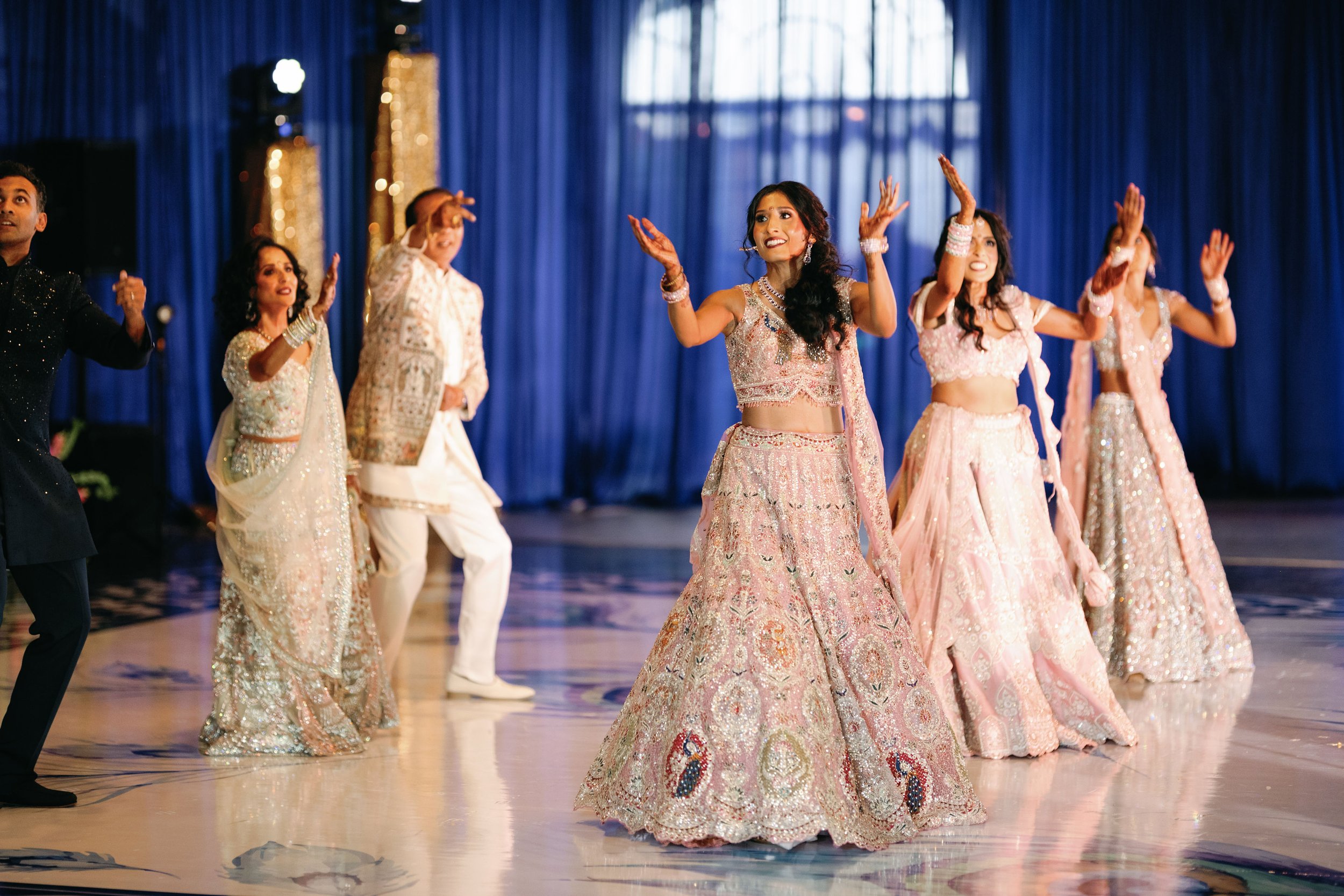 Group of women dressed in traditional, colorful Indian attire performing a dance on stage with a blue curtain background.
