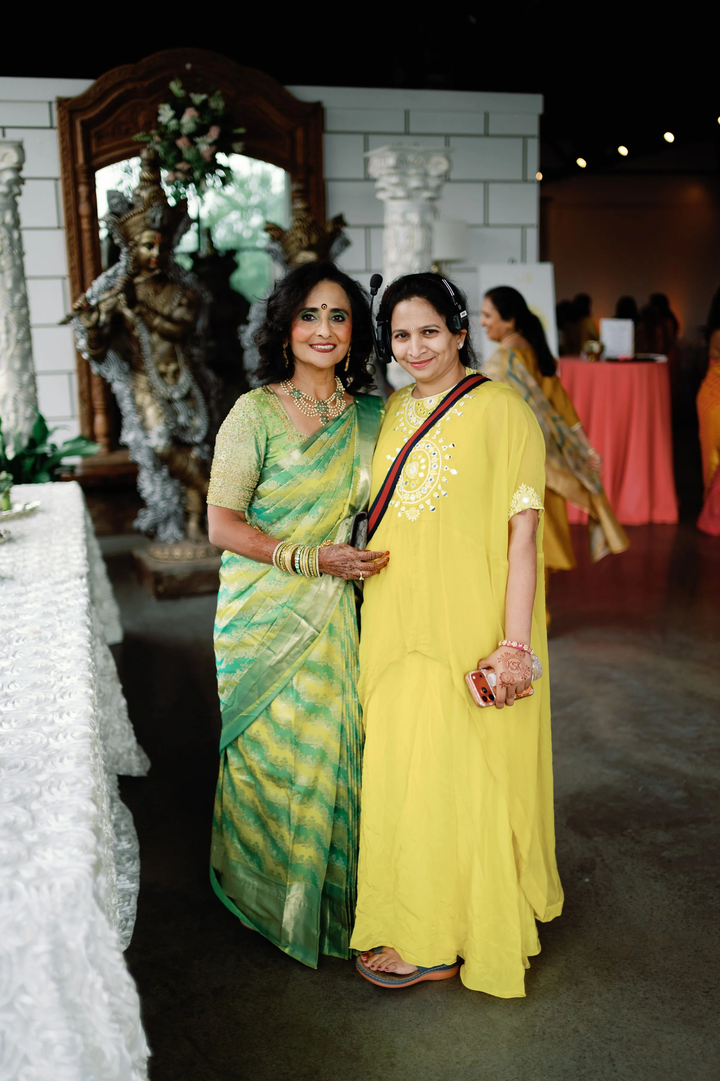 Two women in traditional Indian sarees standing inside a decorated event hall.