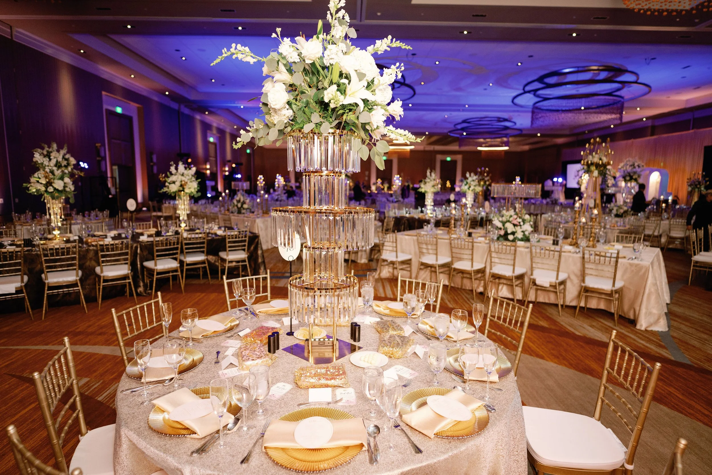 Elegant banquet hall decorated with tall floral centerpieces of white flowers and greenery on round tables with gold chargers, white plates, gold napkins, and glassware, with purple and blue uplighting and ceiling chandeliers.