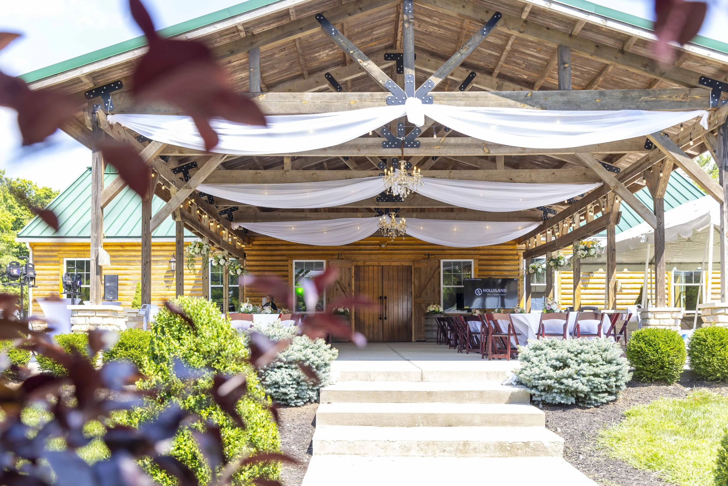 Outdoor wedding reception area with a wooden pavilion decorated with white drapes and chandeliers, surrounded by greenery and flowers.