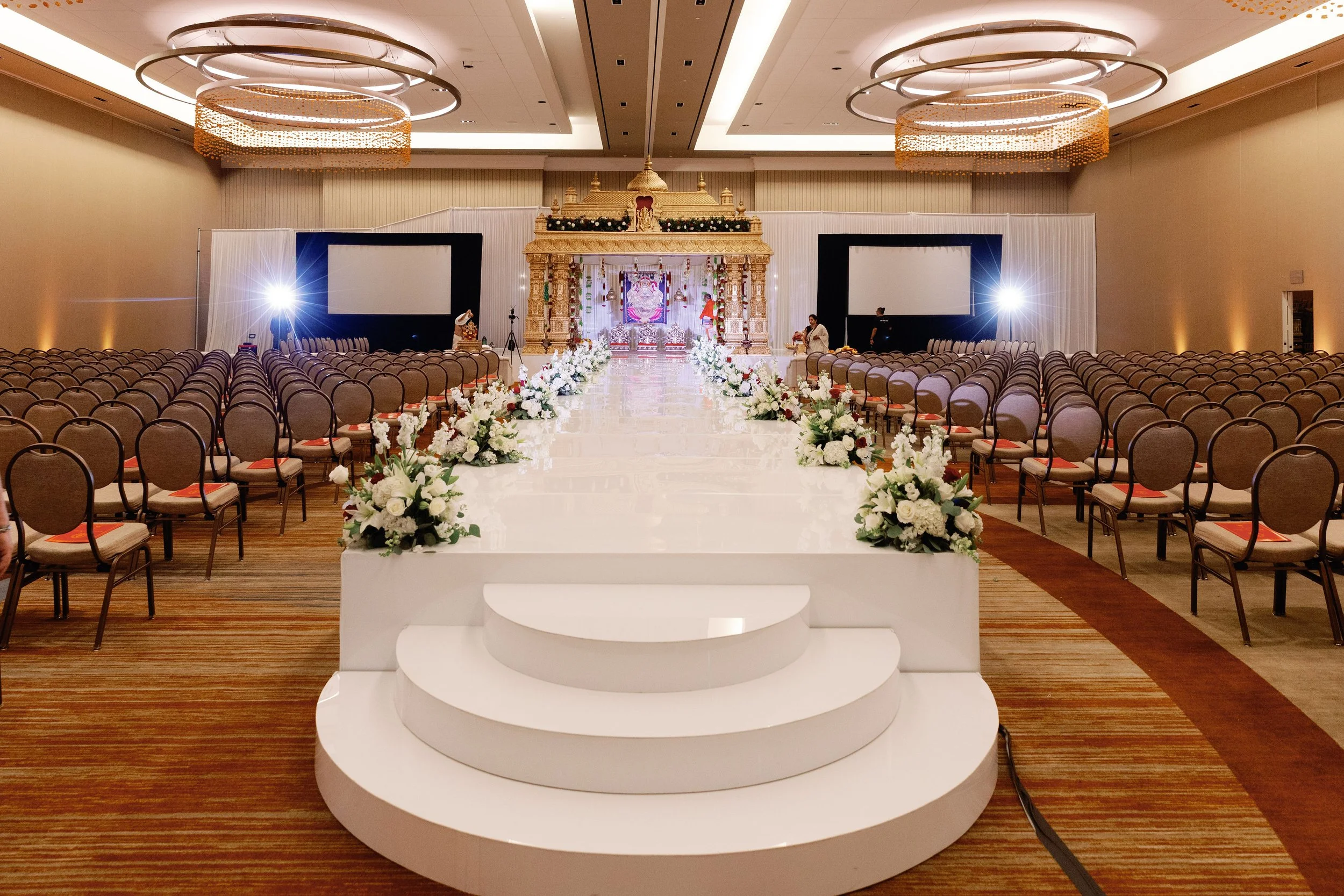 An elegant wedding stage with floral decorations, two large projection screens on each side, and a star-shaped lighting setup. The stage features a decorated golden temple-like structure with traditional Indian elements.