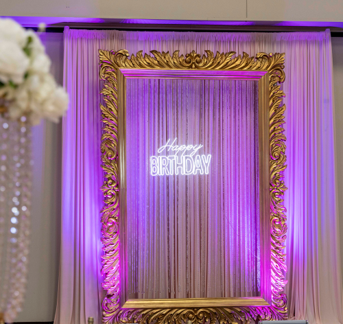 Decorative backdrop for a birthday celebration with a neon 'Happy Birthday' sign, purple lighting, ornate gold frame, and curtains.