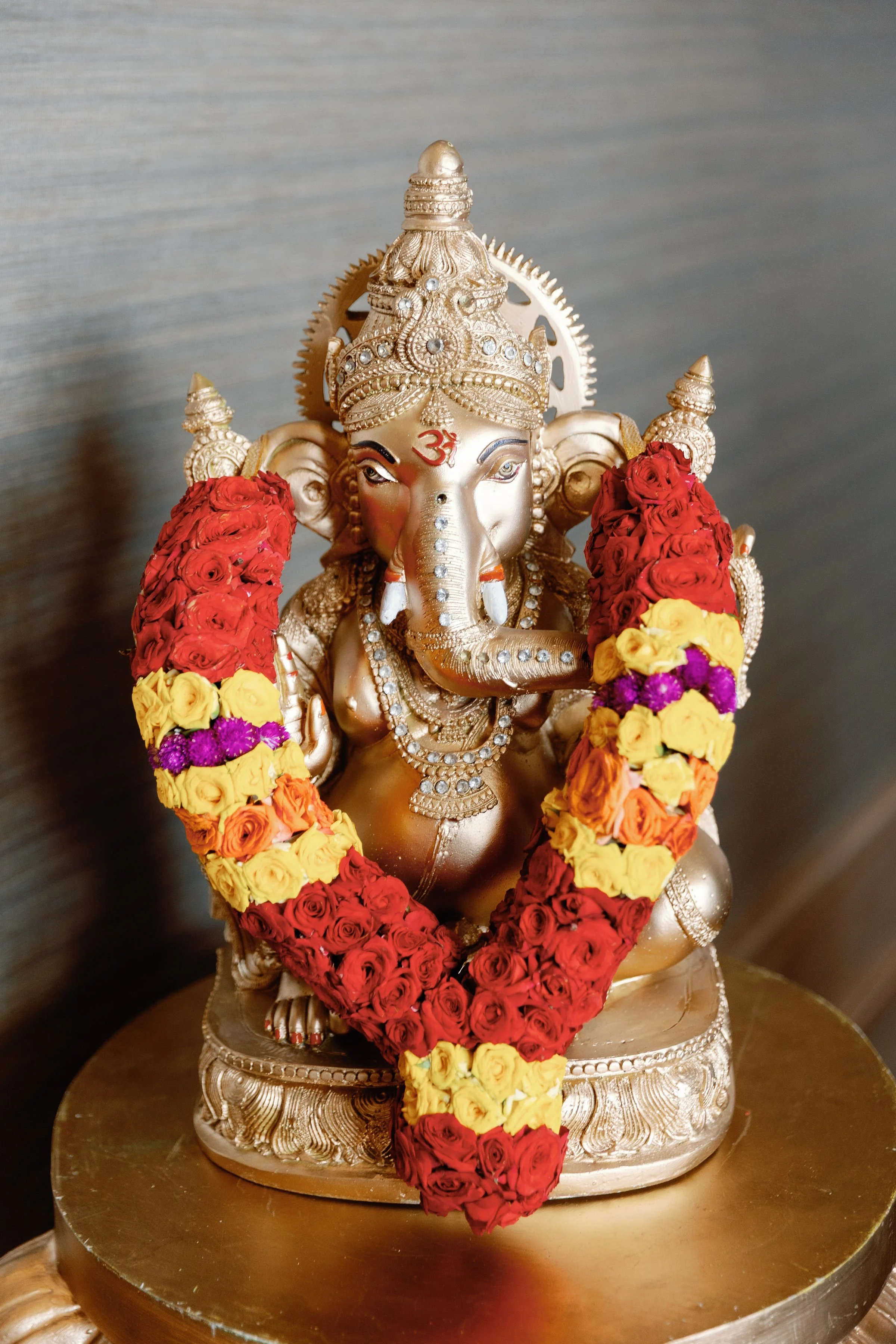 A decorative brass sculpture of Lord Ganesha adorned with a garland of red, yellow, and purple flowers.