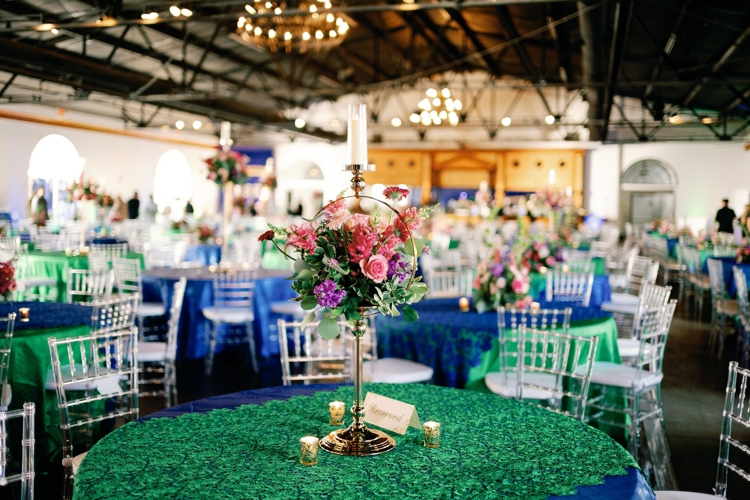 A banquet hall set for a celebration with round tables covered in green and blue tablecloths, decorated with floral centerpieces featuring pink, purple, and red flowers, and surrounded by clear acrylic chairs. A small reserved sign is on one of the tables, and the background shows people and more decorated tables amid warm lighting and chandeliers.