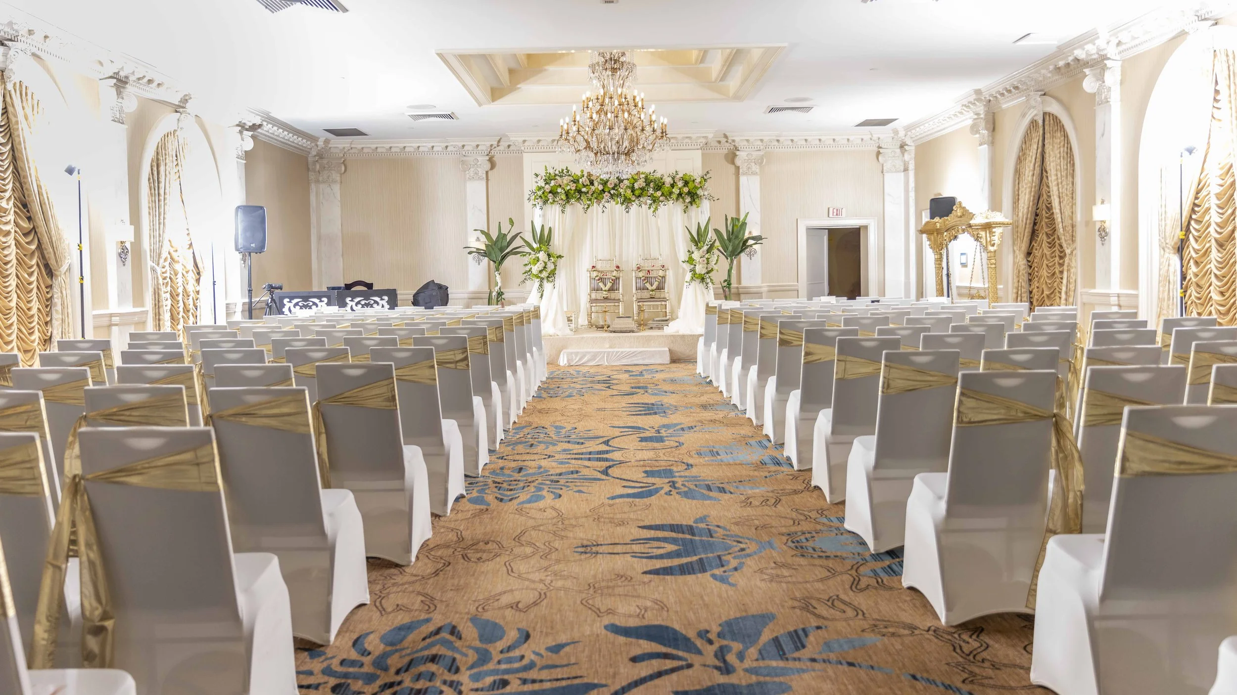 A luxurious wedding ceremony setup with white chairs adorned with gold sashes, arranged in rows facing a decorated altar with floral arrangements and drapery, in an elegant ballroom with chandeliers and ornate details.
