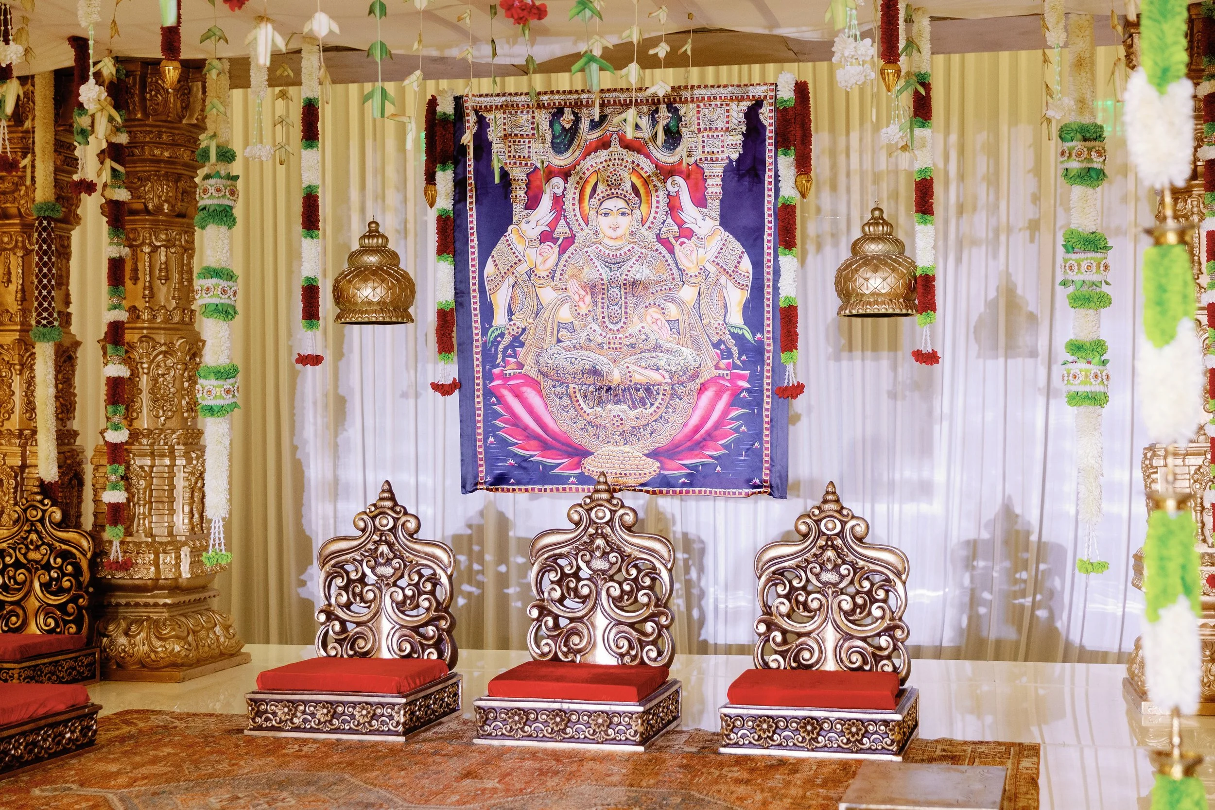 Decorated altar with a large colorful tapestry of a Hindu goddess, surrounded by ornate wooden chairs, hanging decorations, and flowers.