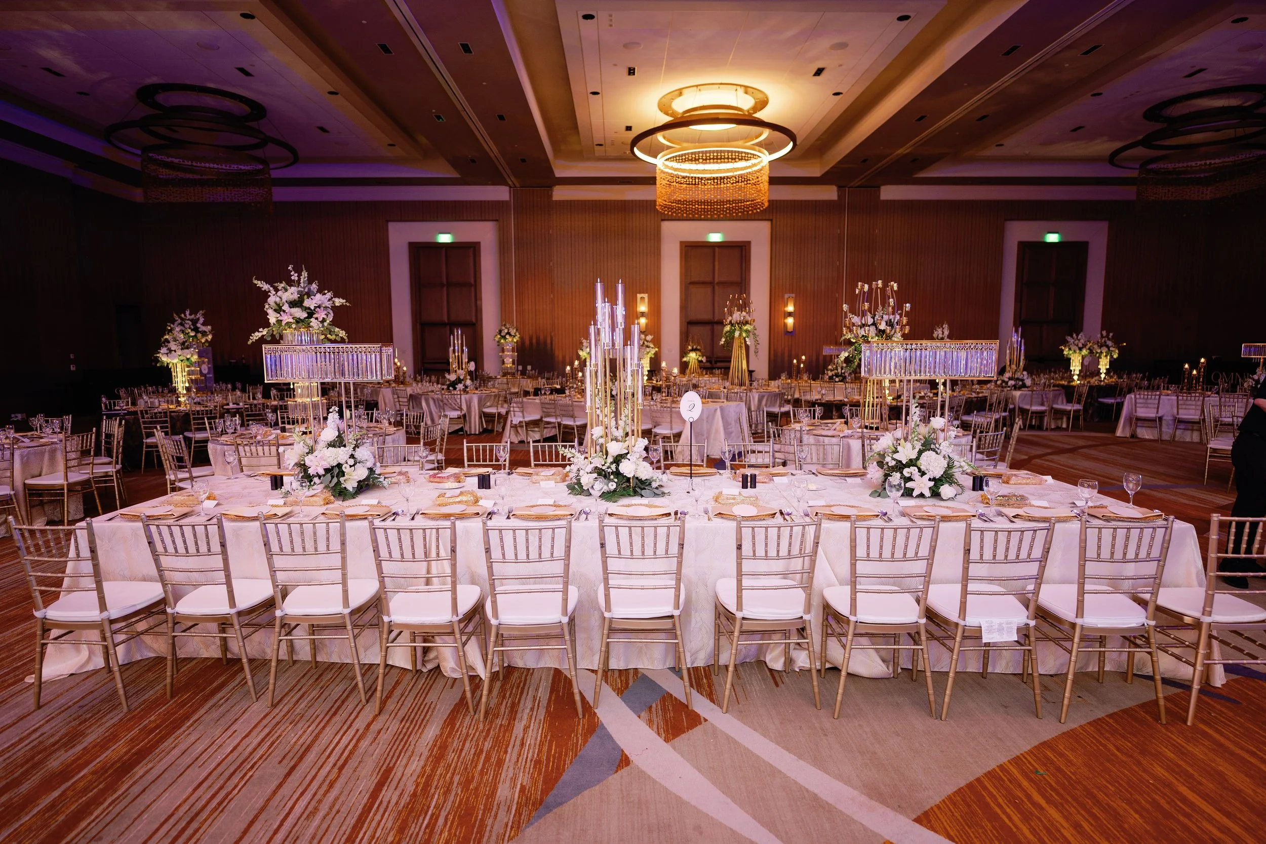 Elegant banquet hall decorated with floral centerpieces, candles, and gold chairs, set up for a formal event or wedding reception.