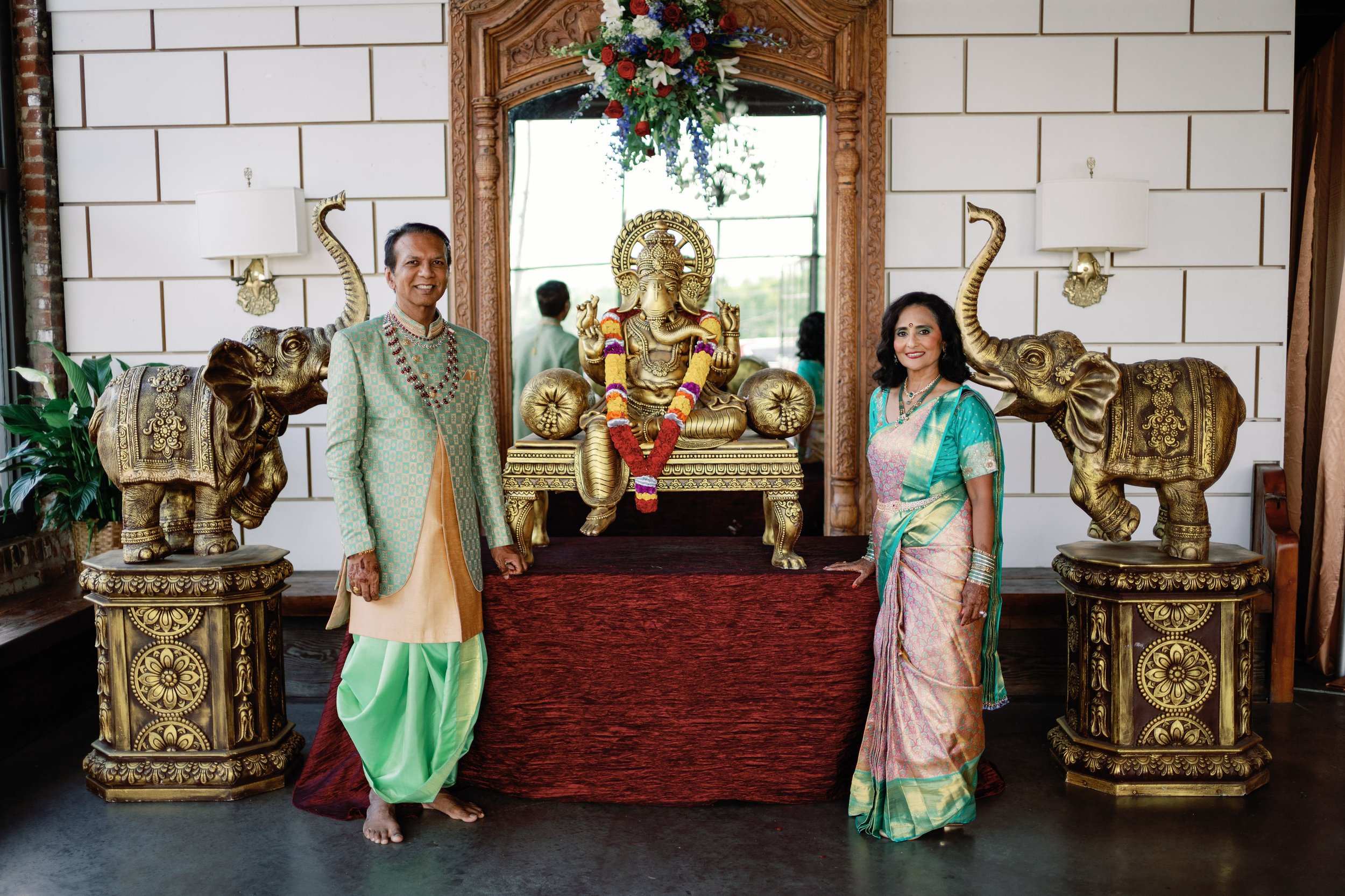 An Indian couple dressed in traditional attire standing beside a golden Ganesha idol decorated with flower garlands, flanked by two ornate elephant statues, in a decorated indoor setting.