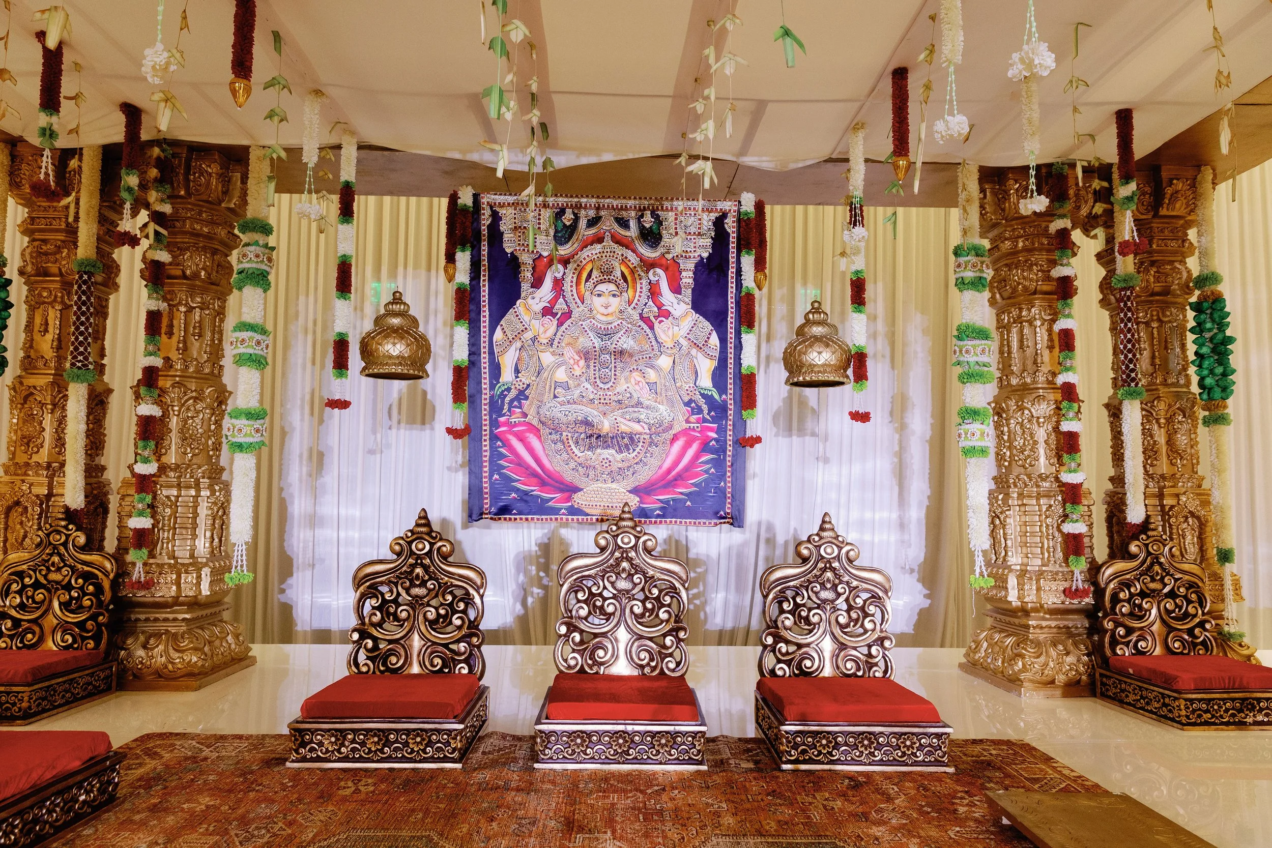 An ornate seating area with carved wooden chairs and cushions, decorated with religious artwork and floral garlands, likely part of a Hindu temple or ceremonial space.