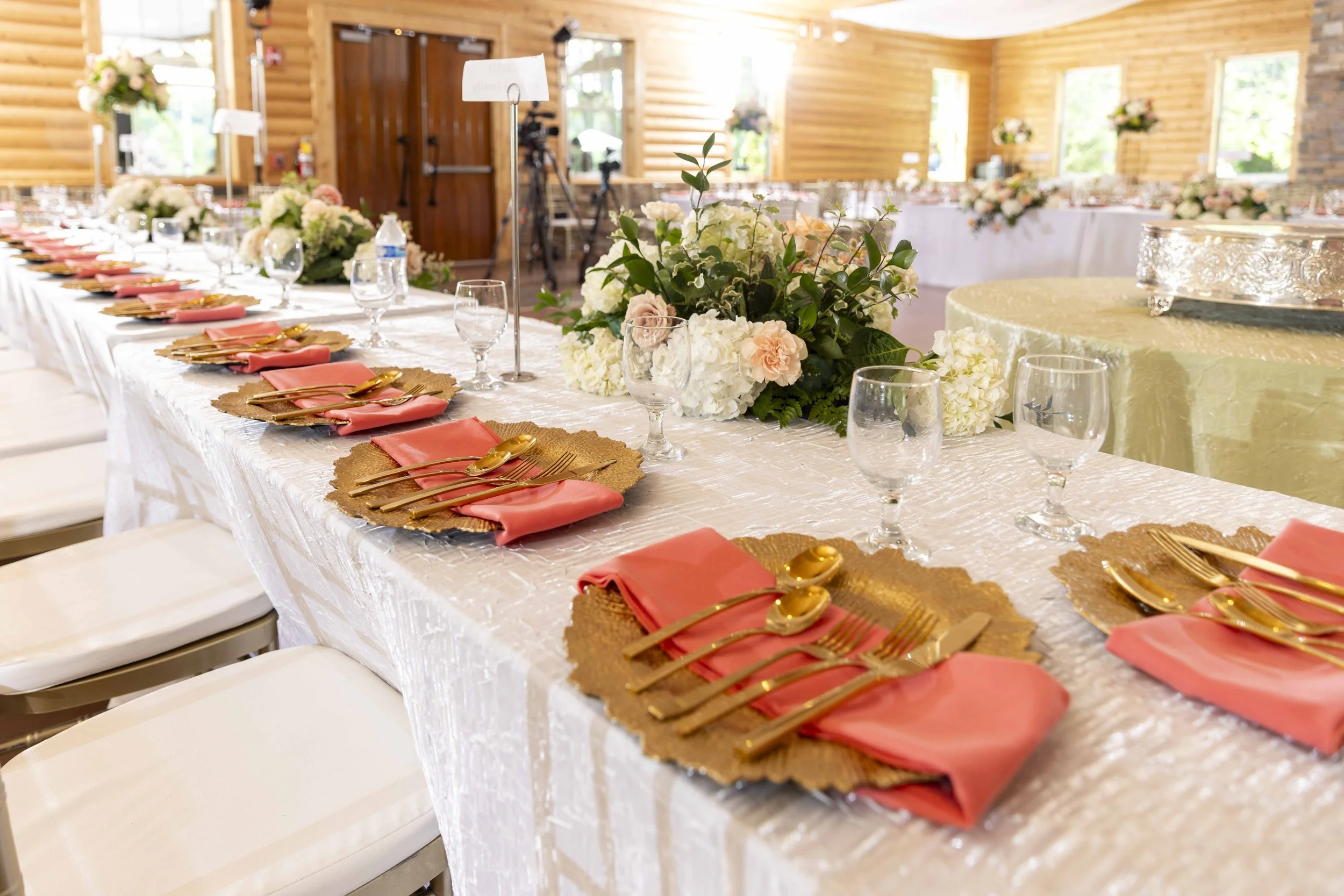 A long banquet table set for a formal event in a wooden hall, decorated with floral centerpieces, pink napkins, gold utensils, and glassware, with a cake in the background.