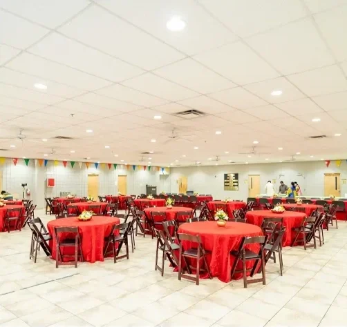 Restaurant or banquet hall with round tables covered with red tablecloths, decorated with floral centerpieces, surrounded by black chairs, and colorful pennant banners hanging from the ceiling.